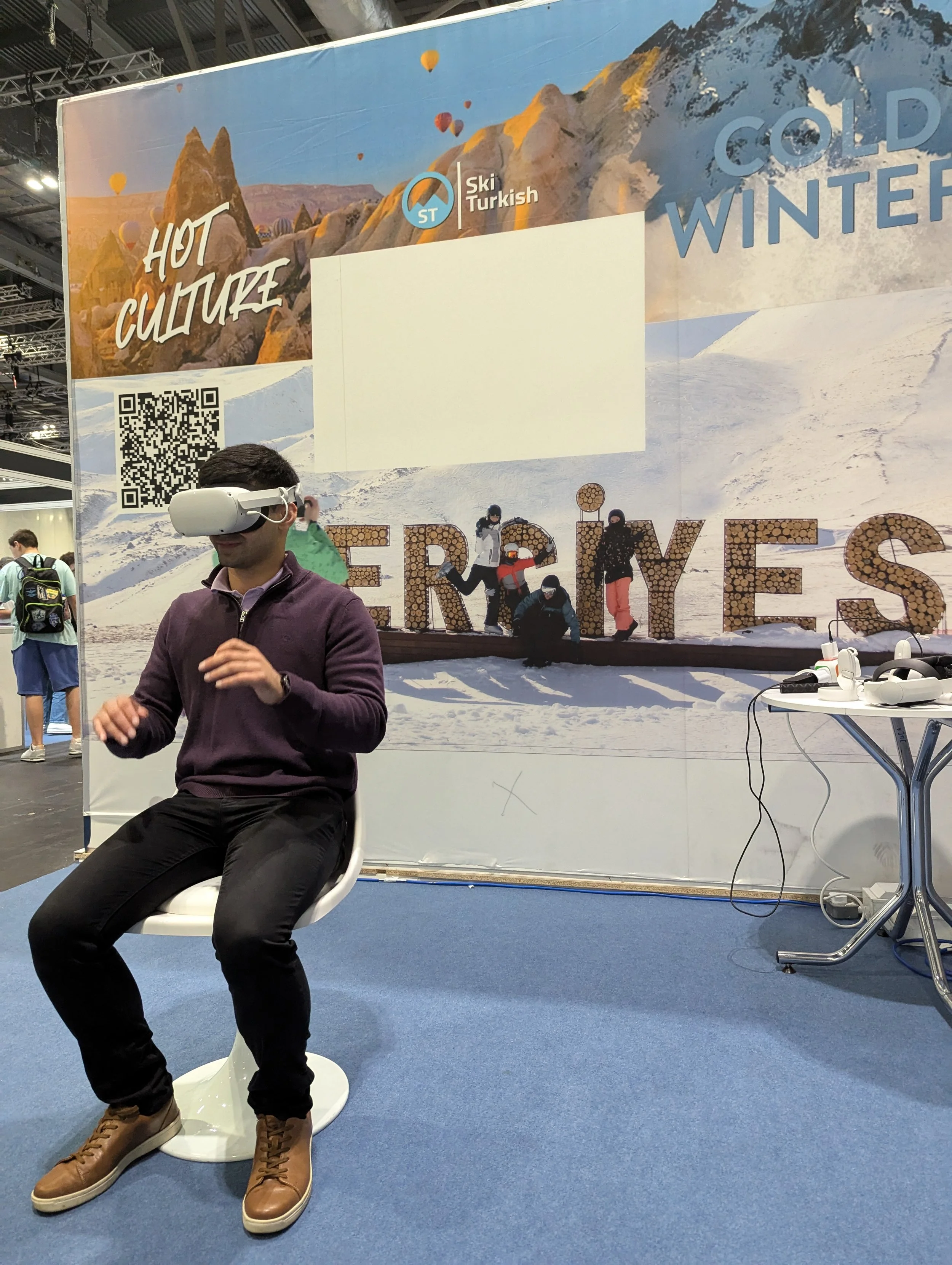 A man sitting on a white chair wearing a virtual reality headset at an indoor event with a large colorful poster in the background promoting winter tourism in Turkey, featuring snowy mountains, hot air balloons, and hikers.