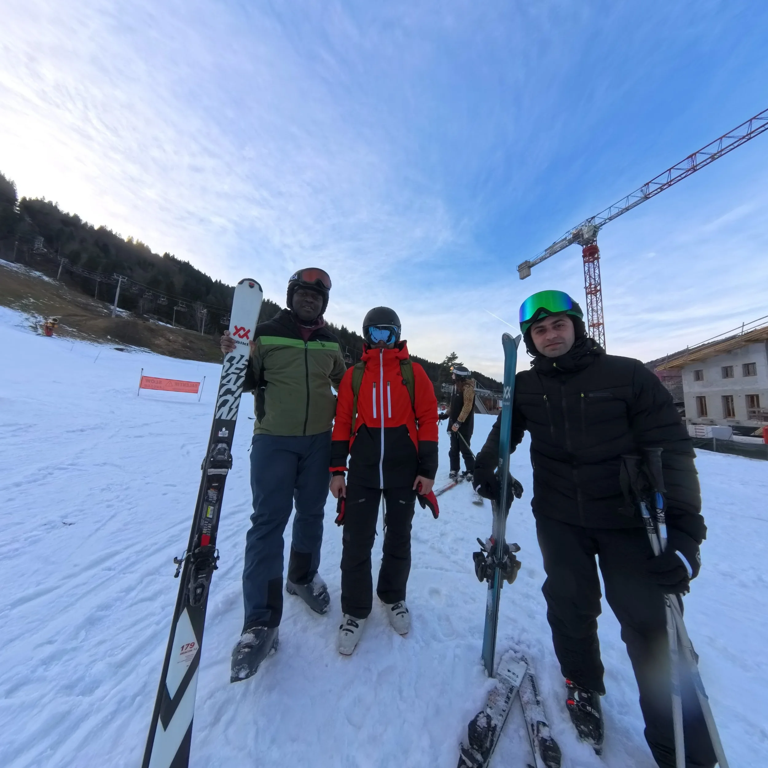 Three people wearing ski gear and helmets standing on snow with skis, in a ski resort area with a crane and building in the background.