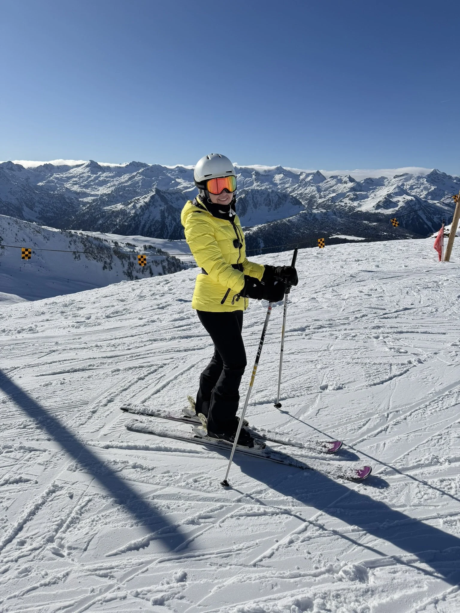 A person in a yellow jacket, black pants, white helmet, and reflective goggles skiing on snowy mountain terrain with a backdrop of snow-capped mountains under a clear blue sky.