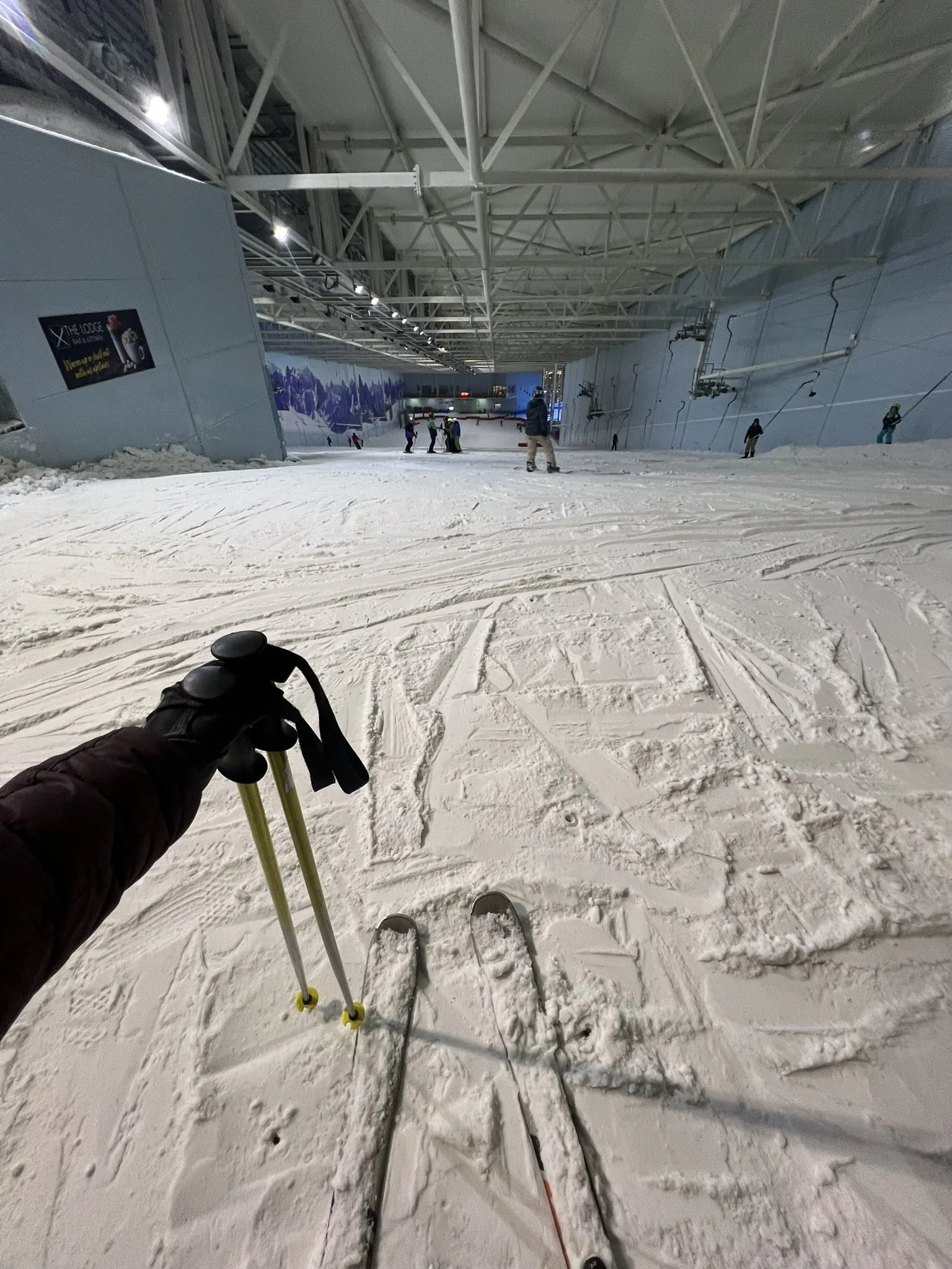 First-person view of a skier holding ski poles inside an underground ski resort, with other skiers and snowboarders in the distance on the snow-covered slope. The ceiling is high and supported by steel beams, with ski lifts visible on the right side.
