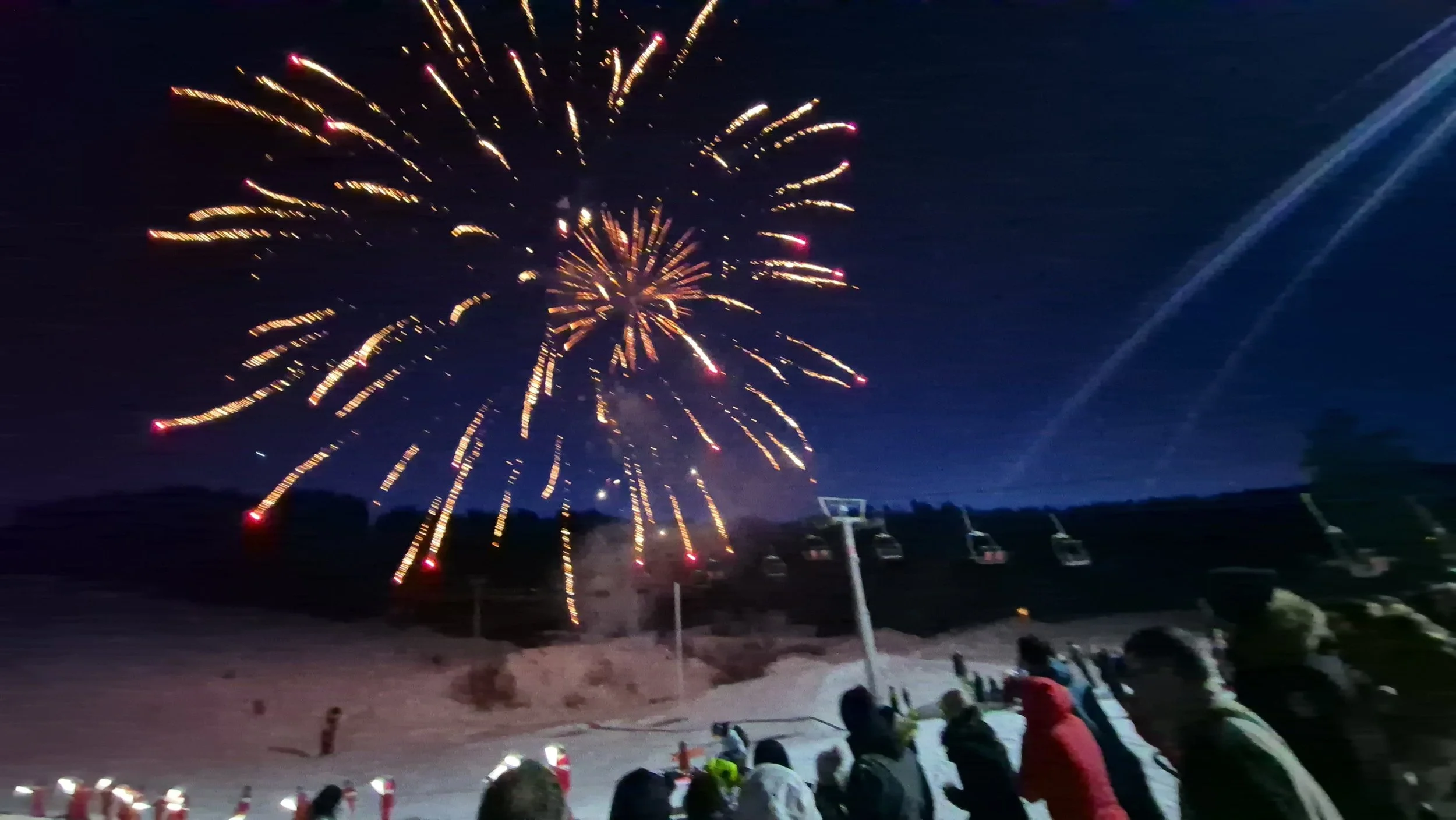 Nighttime scene of fireworks exploding in the sky, with a group of people watching from a snowy slope, some wearing winter jackets.