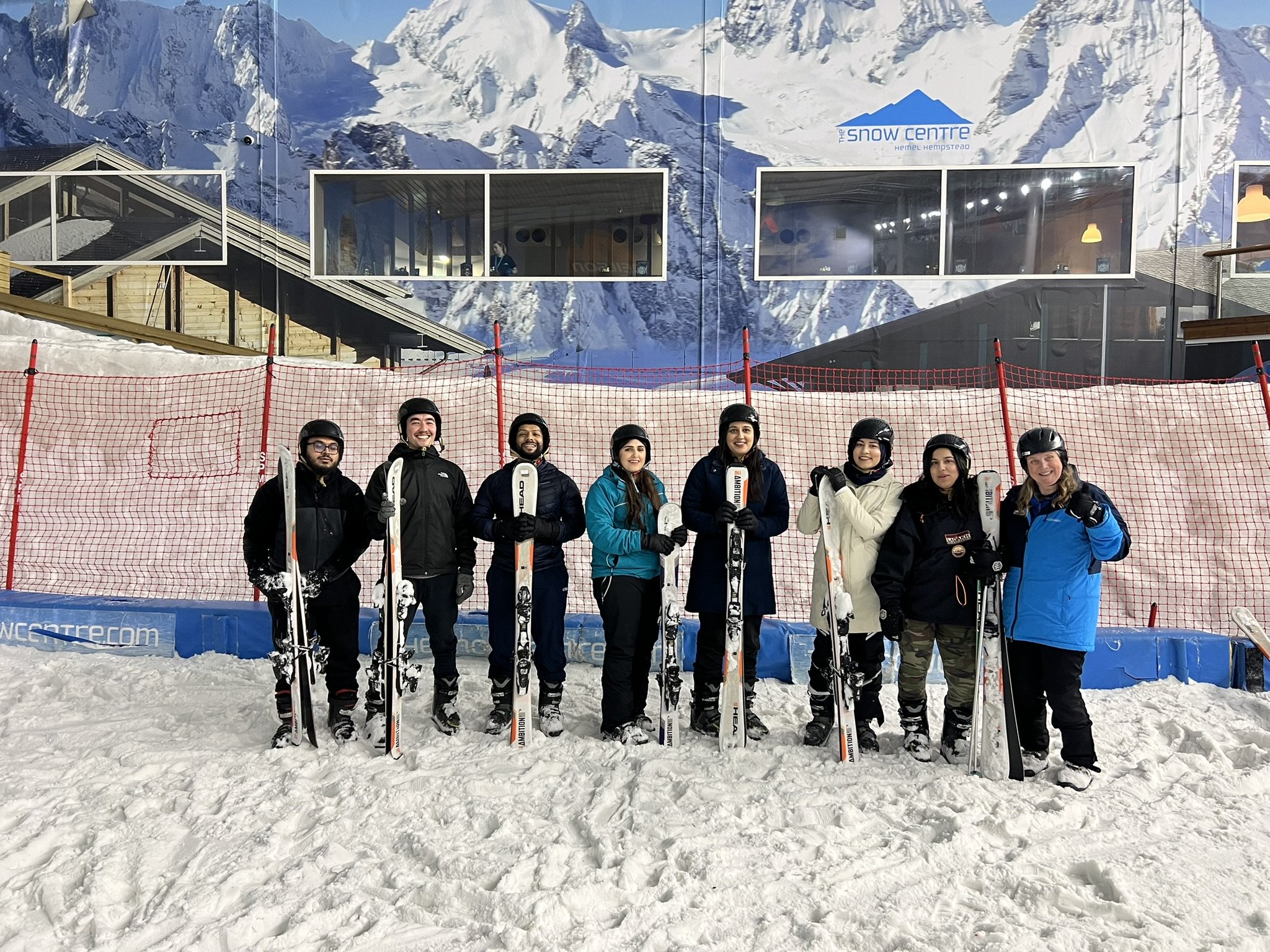 Group of eight people dressed in winter clothing and ski helmets standing with their skis in front of a snow-covered mountain backdrop at a ski resort.