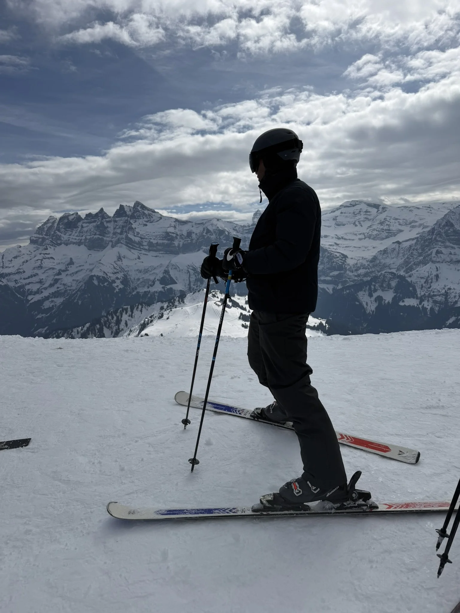A person dressed in winter ski gear, including a helmet, goggles, and gloves, standing on skis with ski poles in hand, on a snowy mountain slope with snow-covered peaks and cloudy sky in the background.