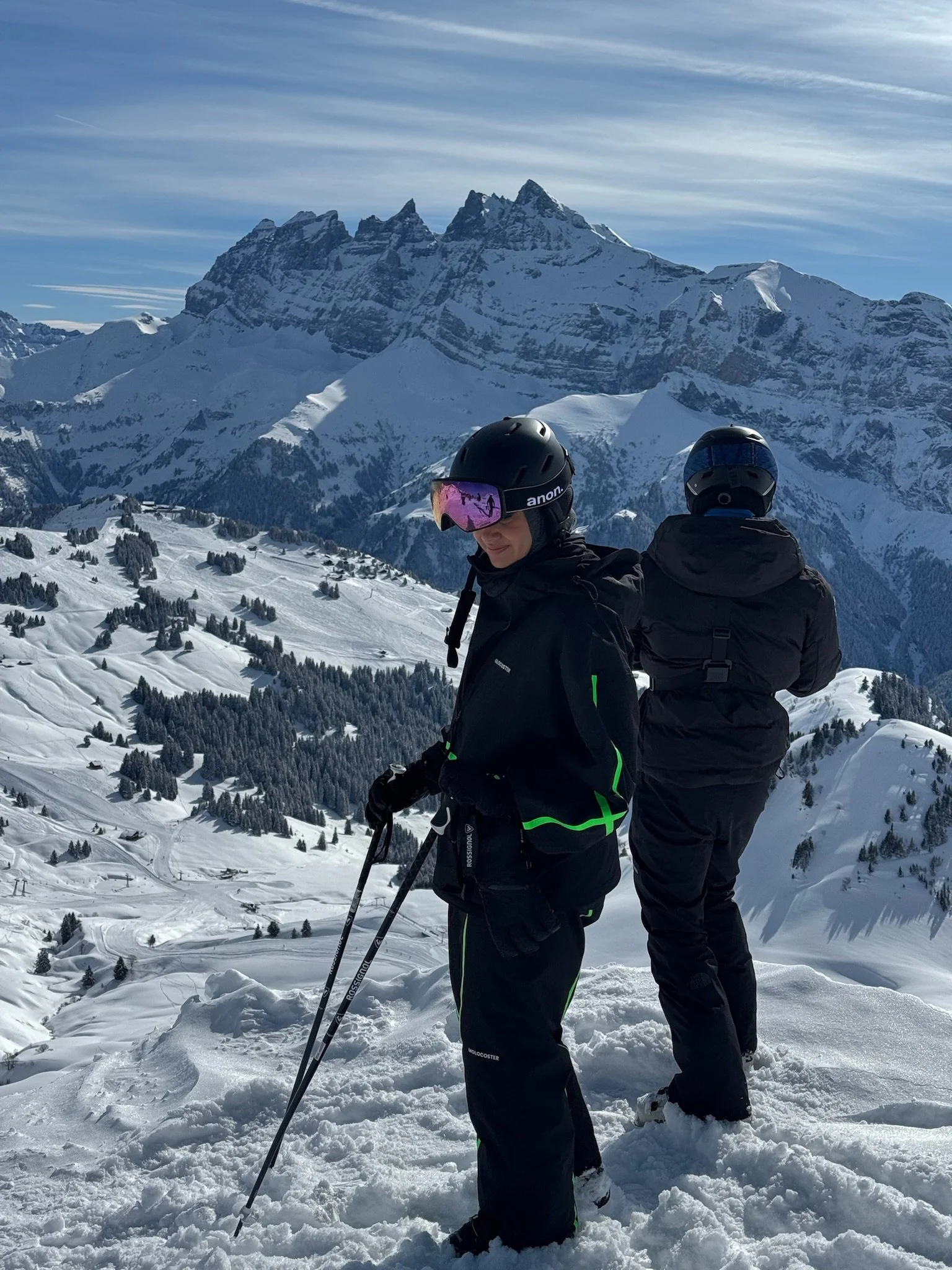 Two people in winter gear with helmets and ski poles standing on snow-covered slope, mountains and a blue sky in the background.