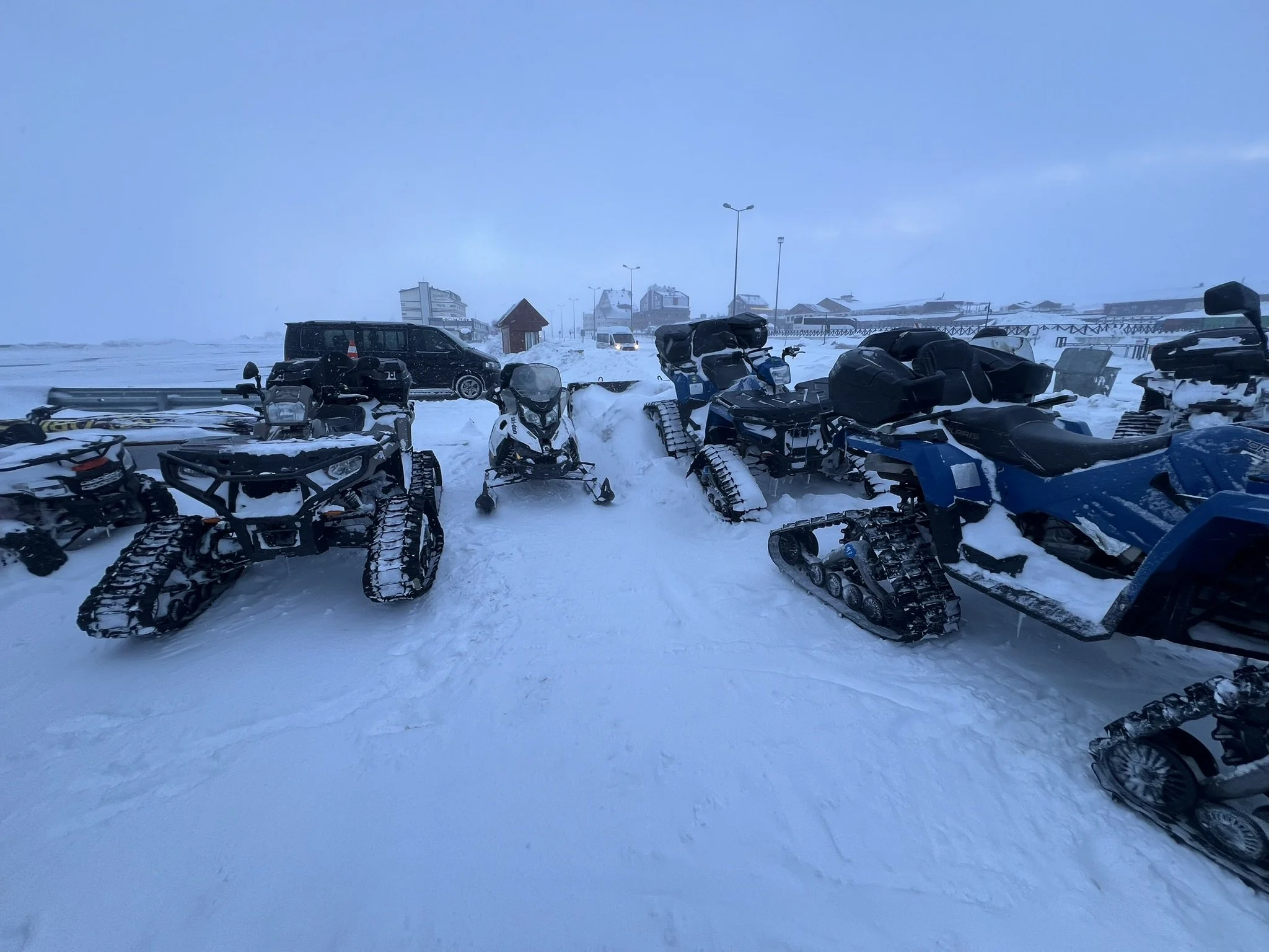 Snow-covered snowmobiles lined up in a snowy landscape with buildings and vehicles in the background.