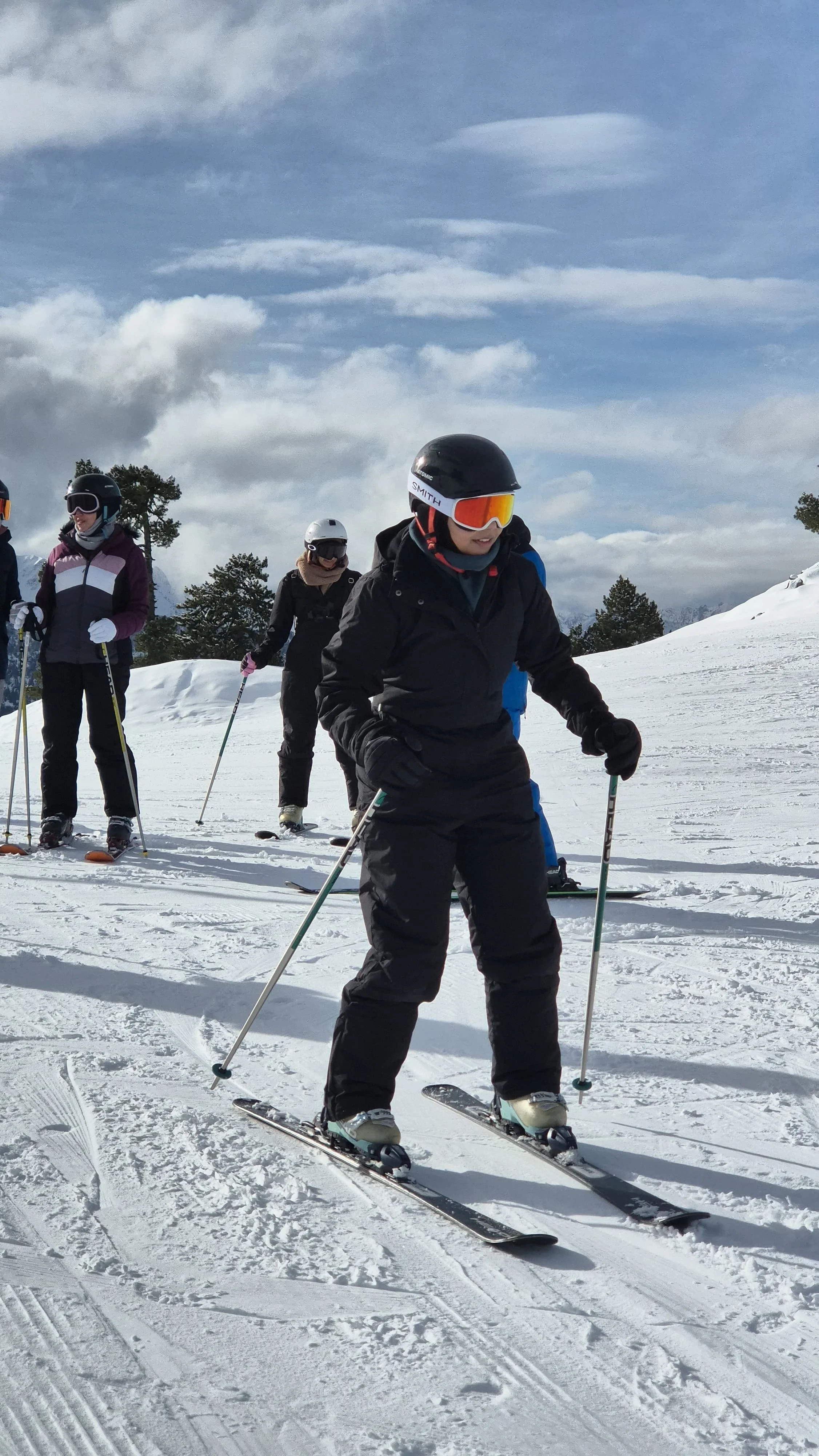 Group of people skiing on snowy mountain, some wearing helmets and goggles, with a cloudy sky above.