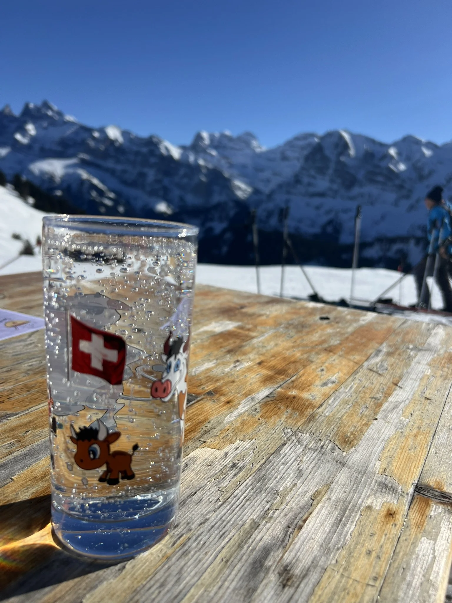 A glass of sparkling water with cartoon horse stickers on a wooden table in a snowy mountain setting with skiers and snow-covered peaks in the background.