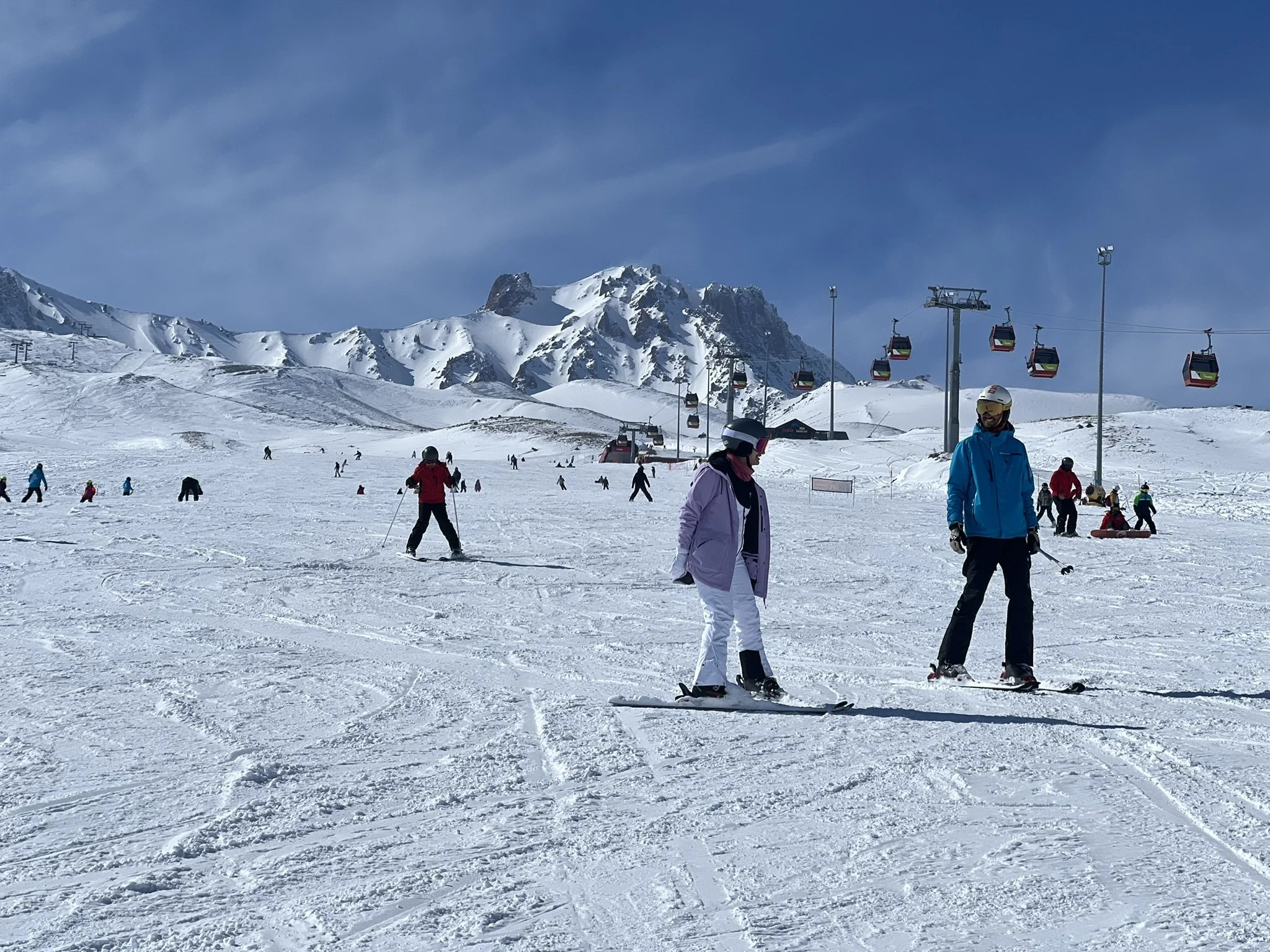 People skiing and snowboarding on a snowy mountain at a ski resort with ski lift chairs, mountain peaks, and a clear blue sky in the background.