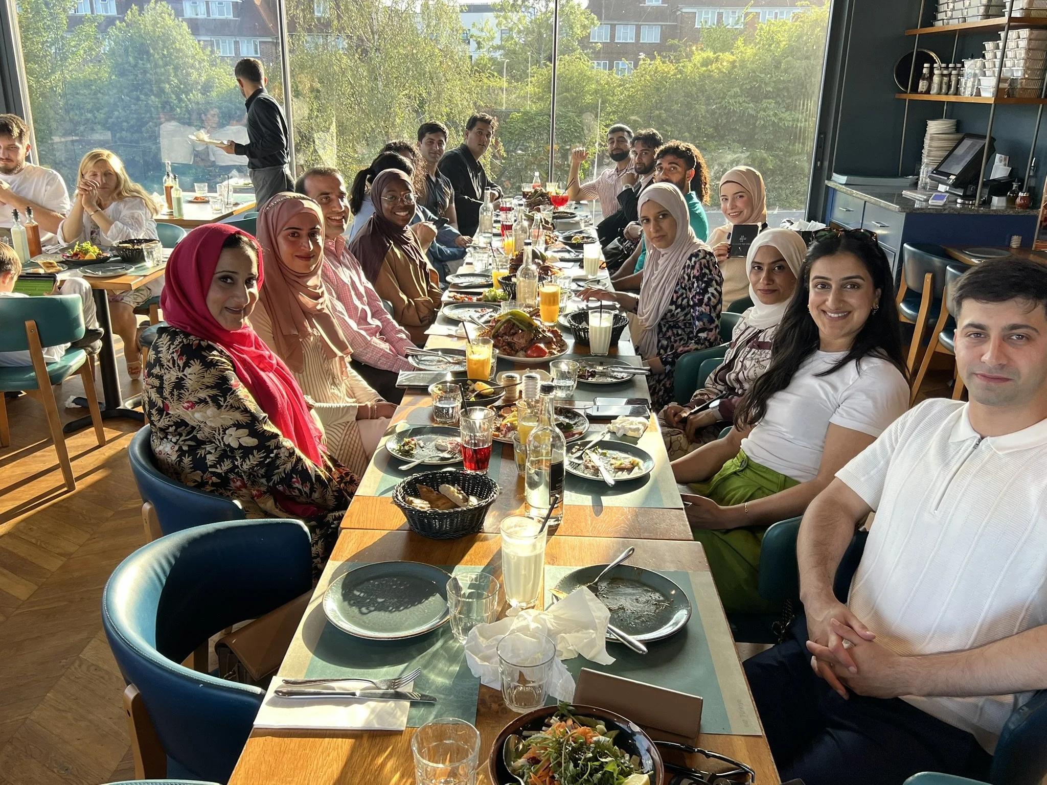 A diverse group of people gathered around a long dining table in a restaurant, enjoying a meal together with plates of food and drinks, with sunlight streaming in through large windows showing outside greenery.