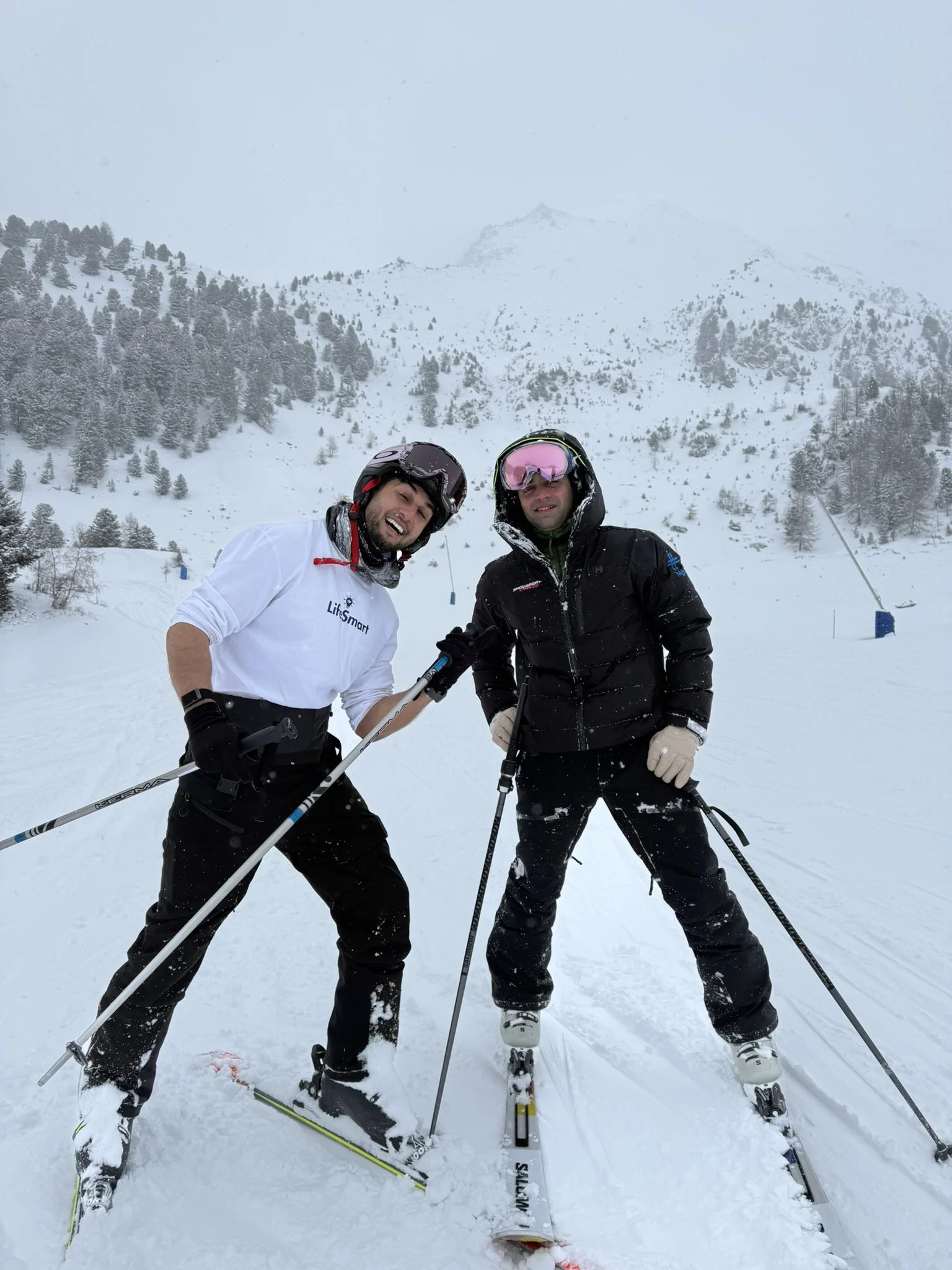 Two men in ski gear posing on a snowy mountain with ski poles in hand, surrounded by snow-covered trees and mountains in the background.