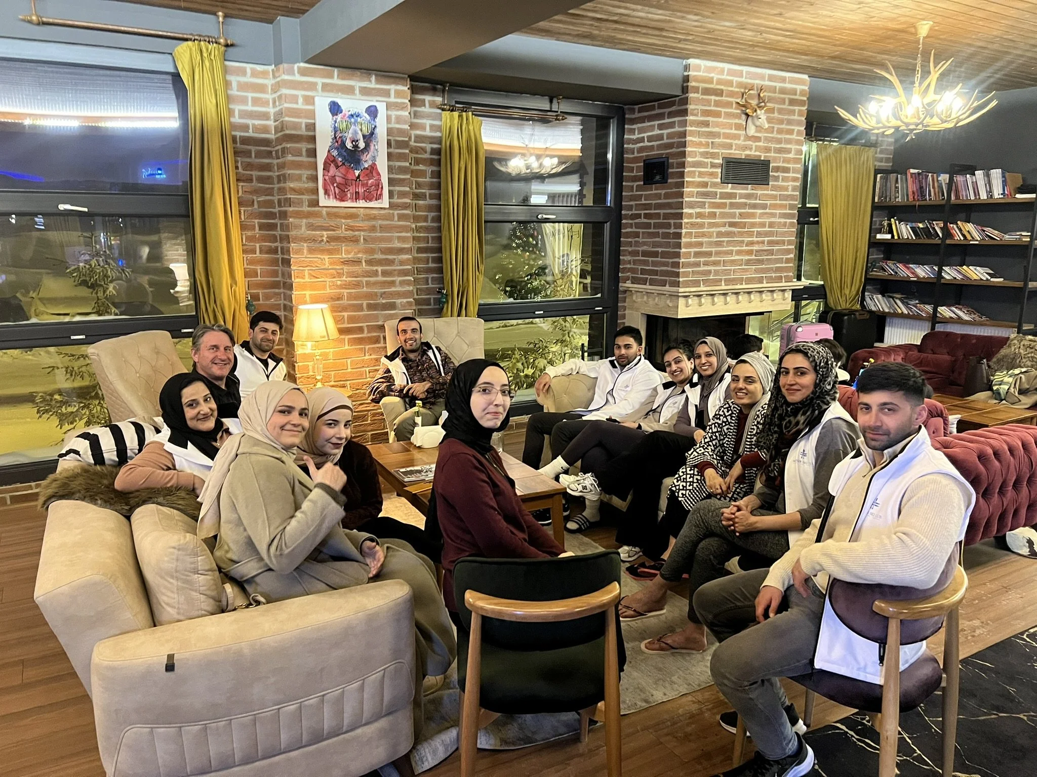 A group of people gathered in a cozy living room, sitting on sofas and chairs, smiling at the camera, with a brick fireplace, bookshelves, and large windows behind them.