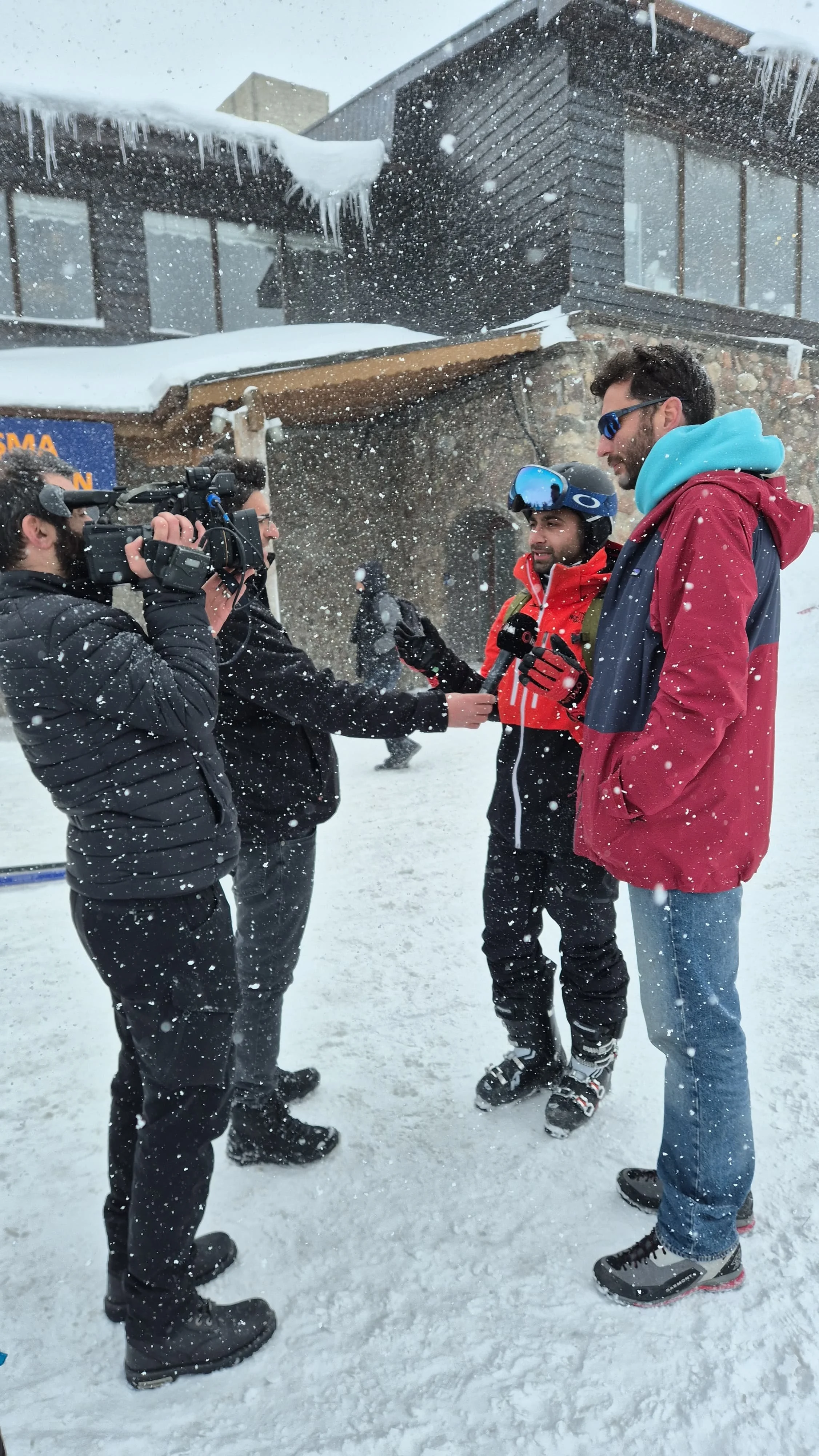 Group of four men in winter clothing and sunglasses, one with a helmet and ski gloves, standing in a snowy outdoor setting being interviewed or recorded by a fifth man with professional camera equipment. Snowfall is visible around them, and there are