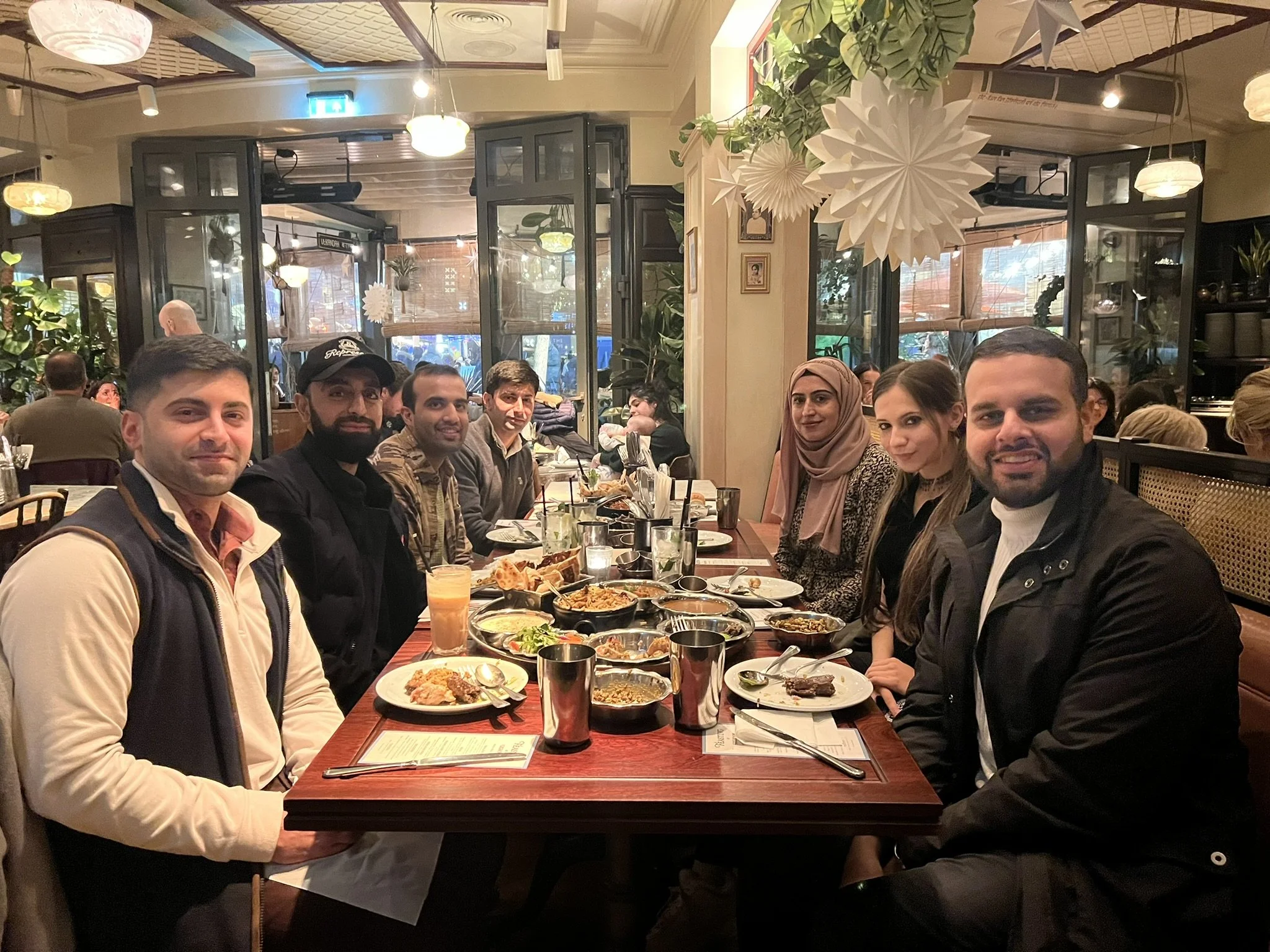Group of seven friends sitting at a dinner table in a cozy restaurant, smiling for the photo, with plates of food, drinks, and utensils on the table, warm lighting, and decorative paper lanterns hanging from the ceiling.