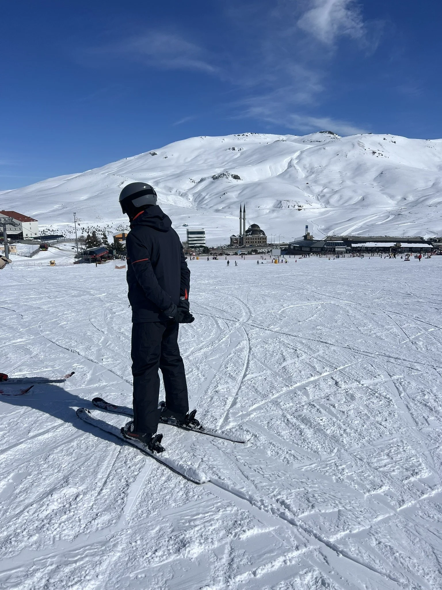 Person standing on snow-covered ground, wearing a black helmet and winter jacket, with ski equipment, with snow-capped mountains, ski slopes, and a blue sky in the background.