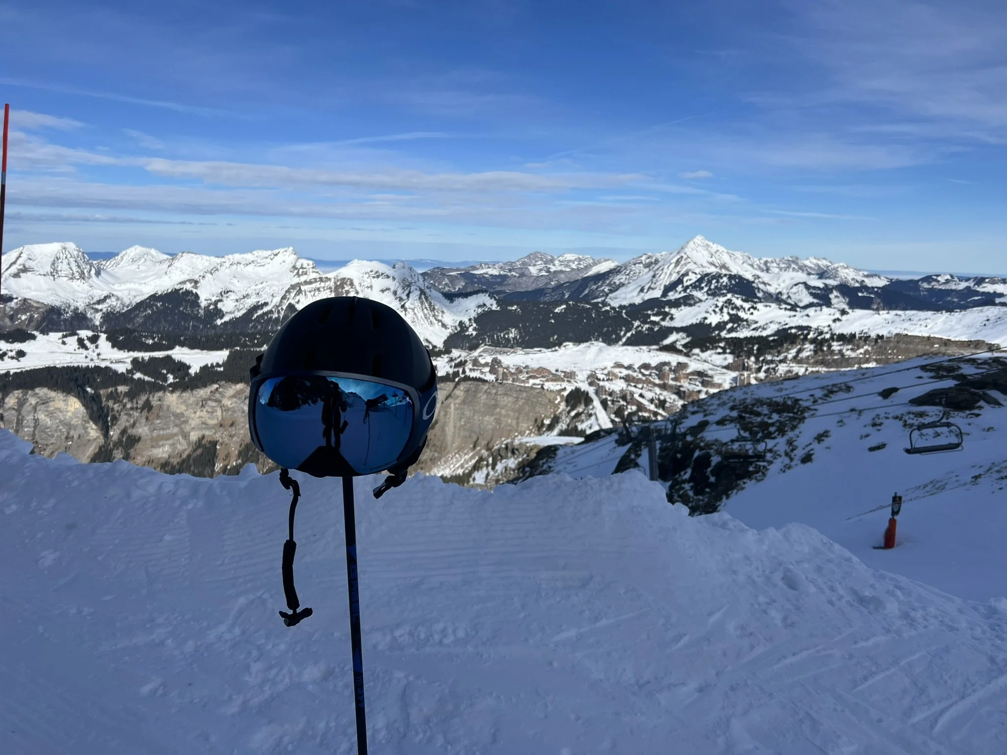 A black ski helmet with blue reflective goggles resting on a ski pole on a snowy mountain slope with snow-capped mountain peaks and blue sky in the background.