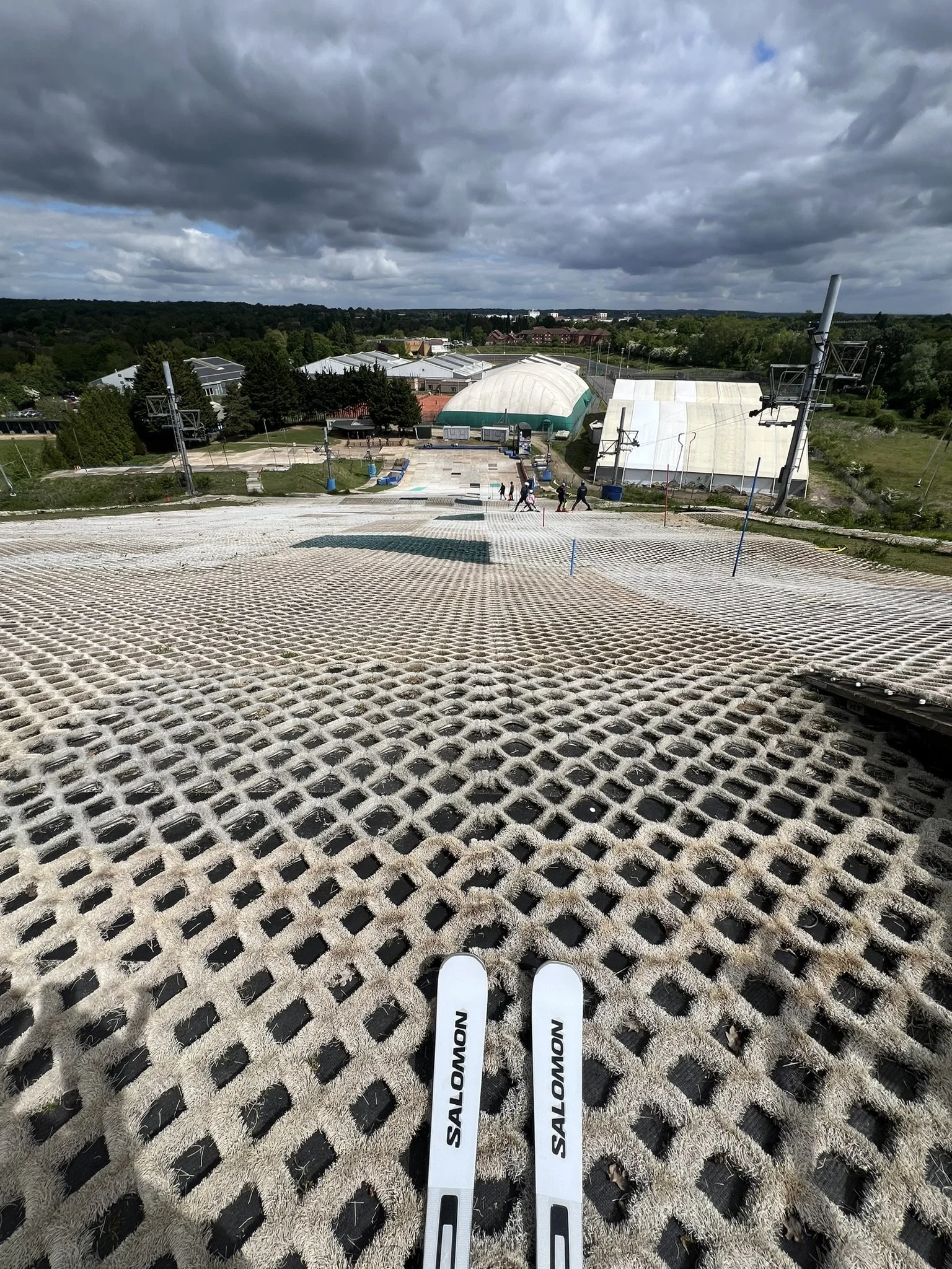 View from the top of a ski slope with Salomon skis at the bottom, and a ski lift in the distance, under a cloudy sky.
