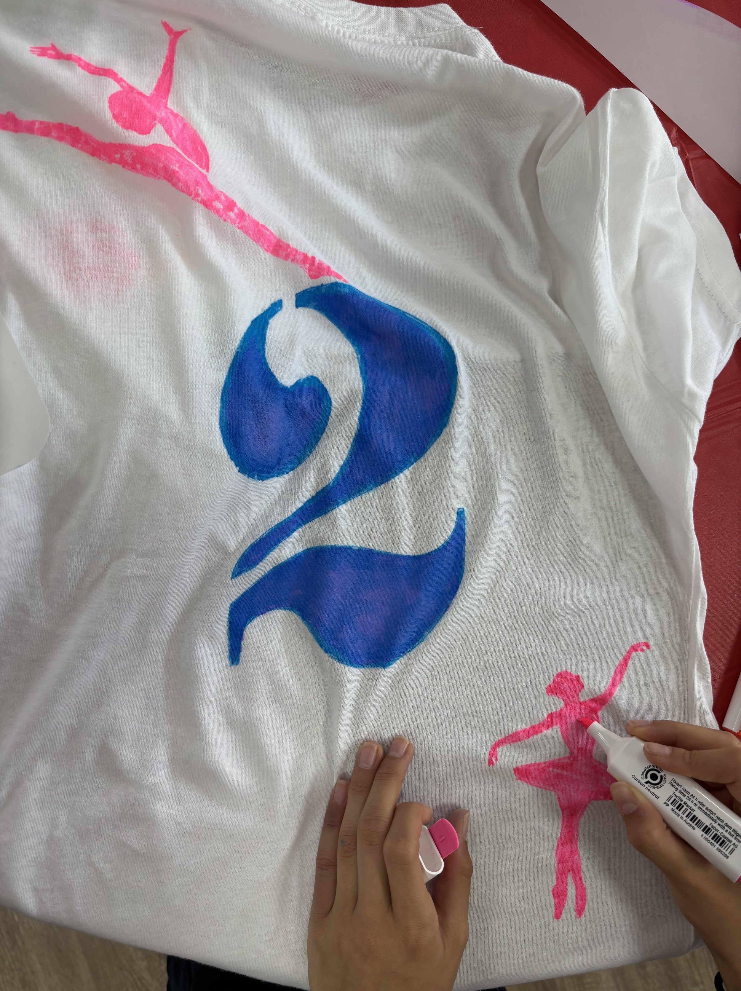 Person drawing pink ballerina and blue number on a white T-shirt with markers.