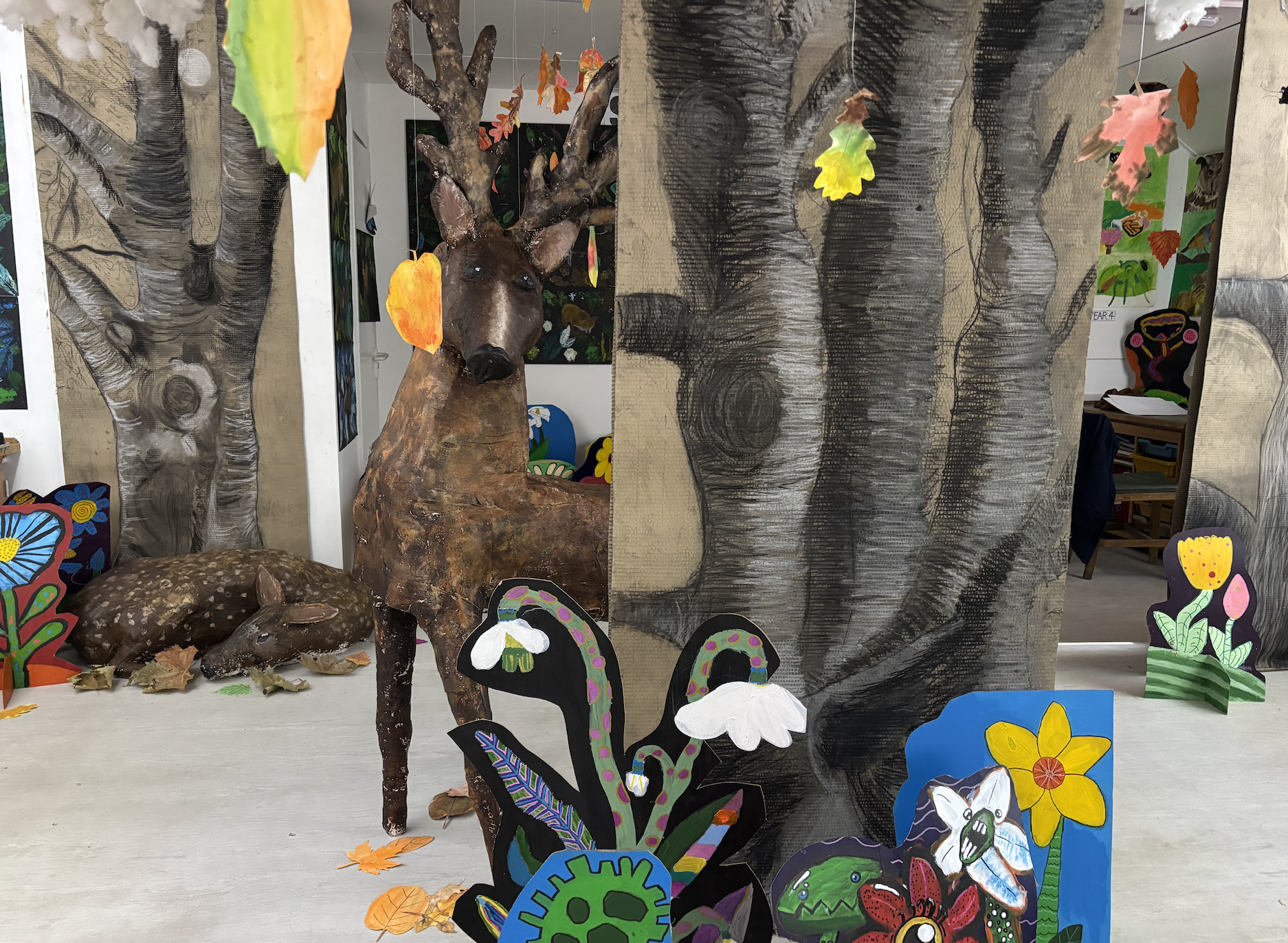 A zoo or nature-themed art display featuring a carved and painted deer with antlers, surrounded by colorful painted cutouts of flowers and insects, with small leaves scattered on the surface.