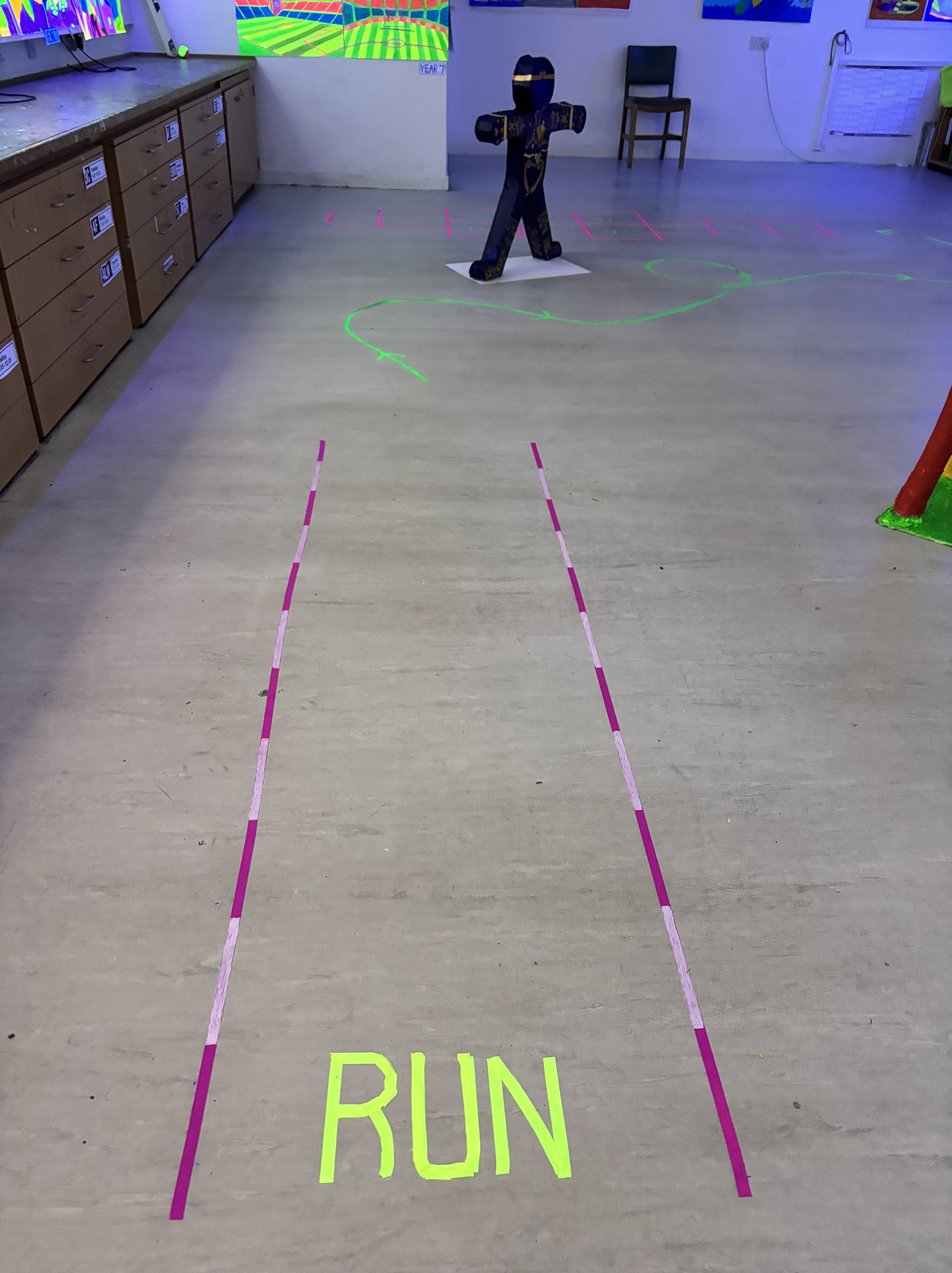 Indoor activity area with glow-in-the-dark markings and a green line guiding a running path, labeled 'RUN'. There is a cardboard cutout of a walking figure and a cross sculpture, with a TV screen in the background showing a virtual sports game.