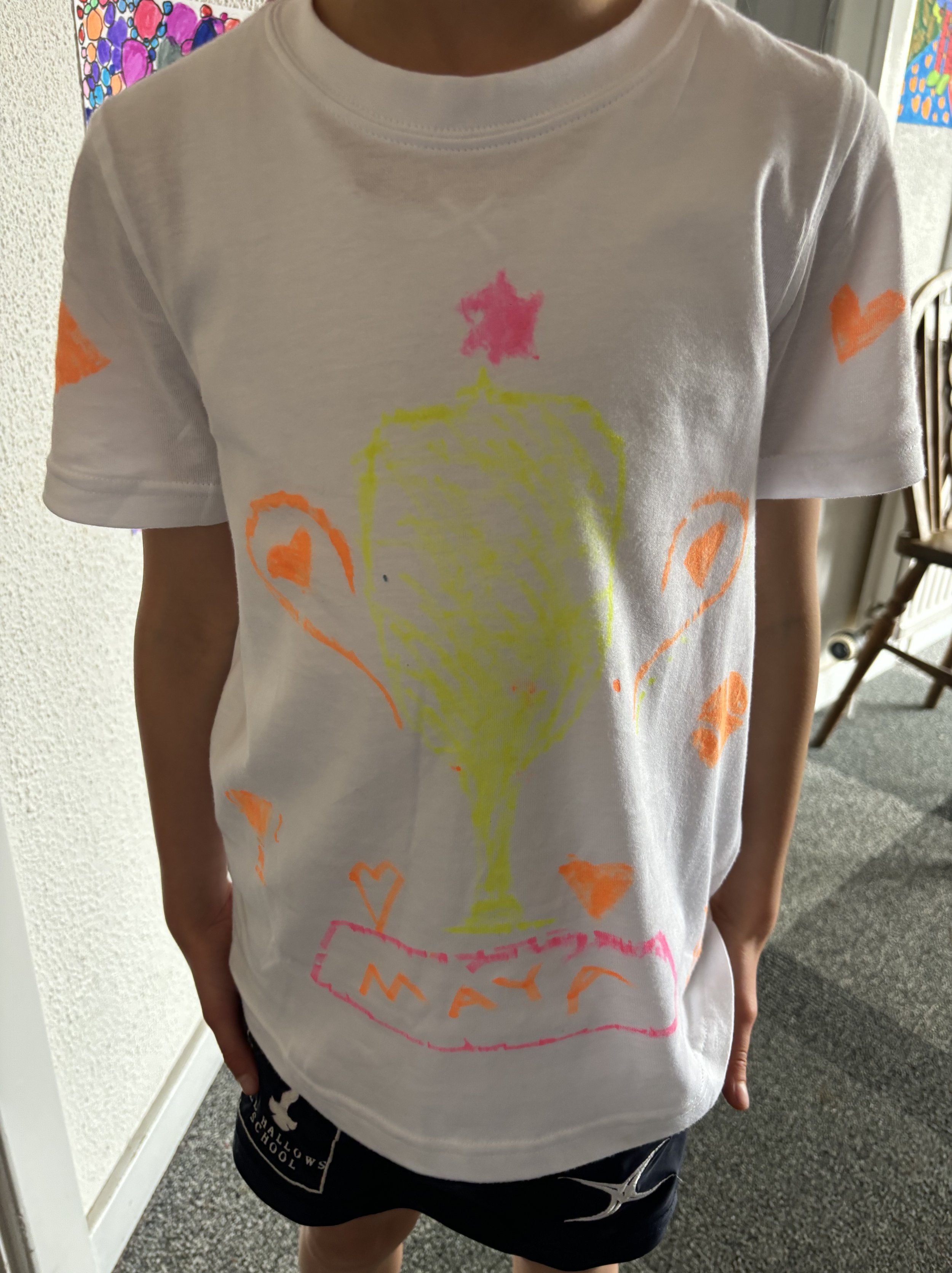 Child wearing a white t-shirt with a colorful, hand-drawn design of a large glass of champagne or wine with a pink star on top, several orange hearts, and the word "MAY" written in orange below the glass.