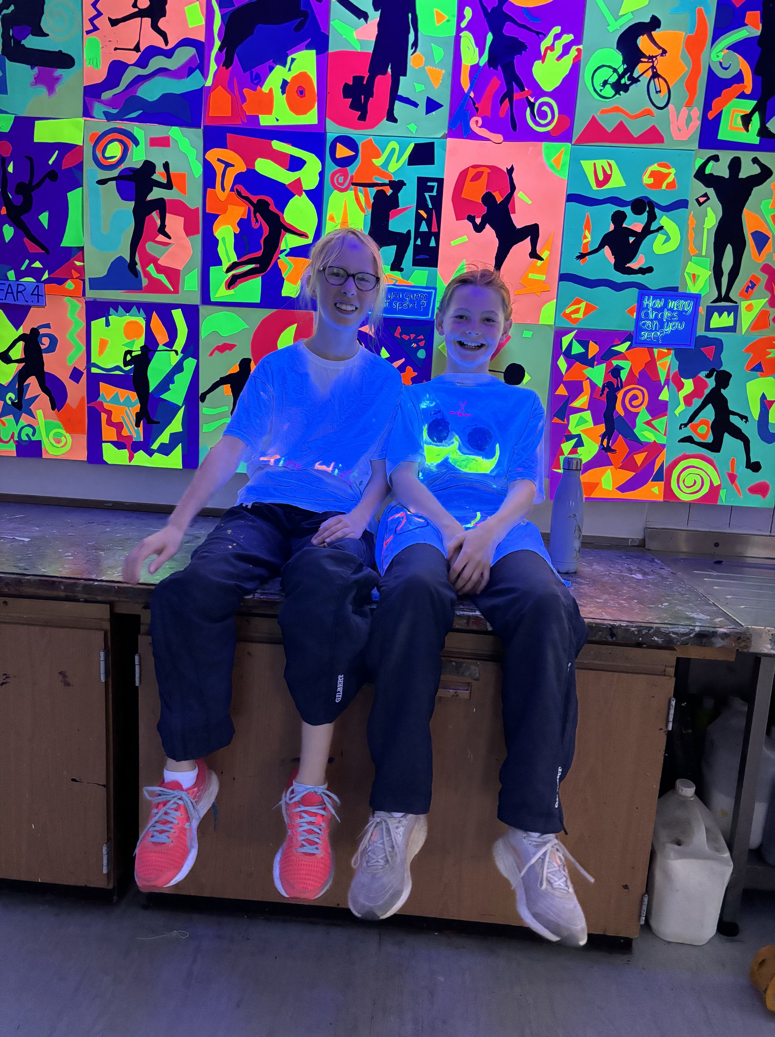 Two young girls sitting on a wooden table in front of a vibrant, neon-colored artwork with silhouettes of people engaging in various sports and activities. One girl is wearing glasses, and both are smiling. The artwork features bright colors and abst
