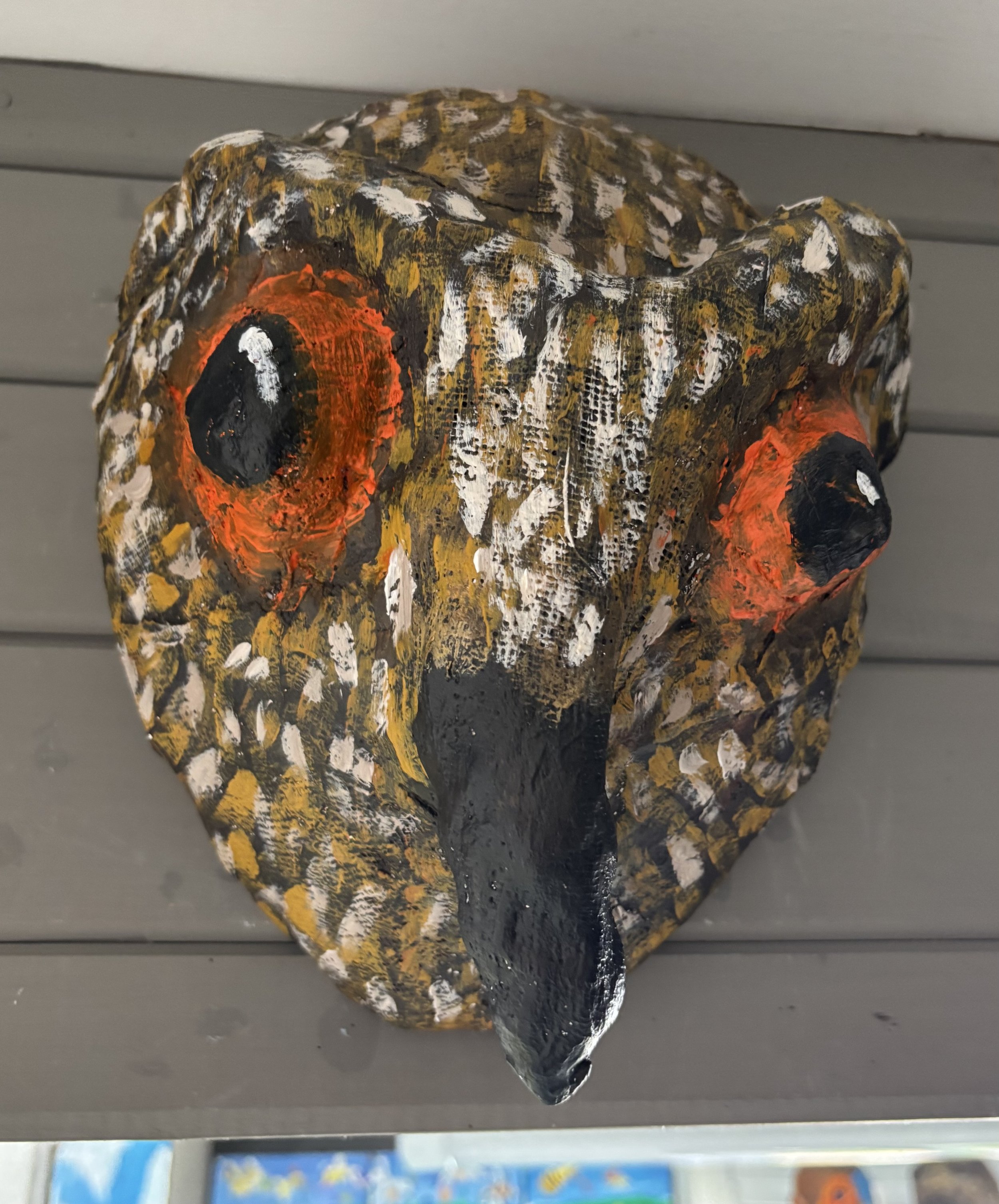 Hand-painted owl mask with prominent eyes, beak, and owl feather pattern, mounted on a wall.