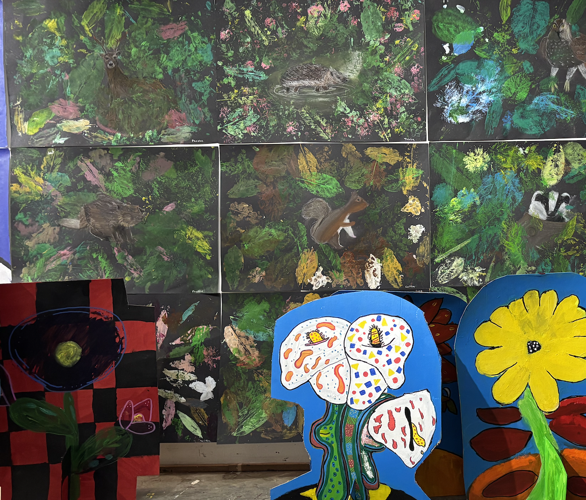 A display of various colorful, child-like paintings. The top section features painted animals like a deer, a raccoon or small mammal in a foliage background. The bottom section includes a painted flower on a blue standing board, another large yellow