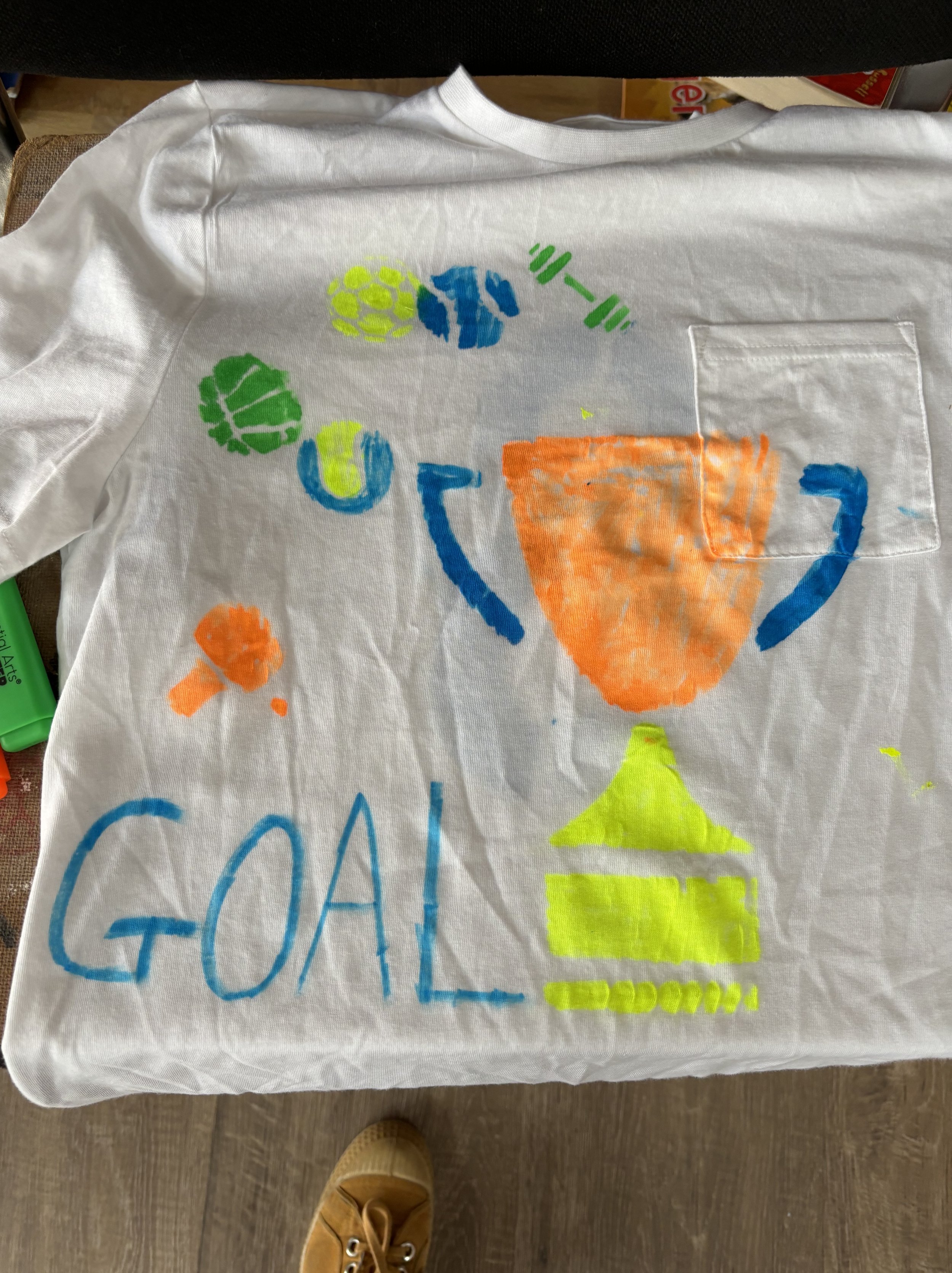 Hand-painted design on a white fabric, featuring a colorful trophy, the word 'GOAL' in blue, and various abstract shapes in blue, green, orange, and yellow.