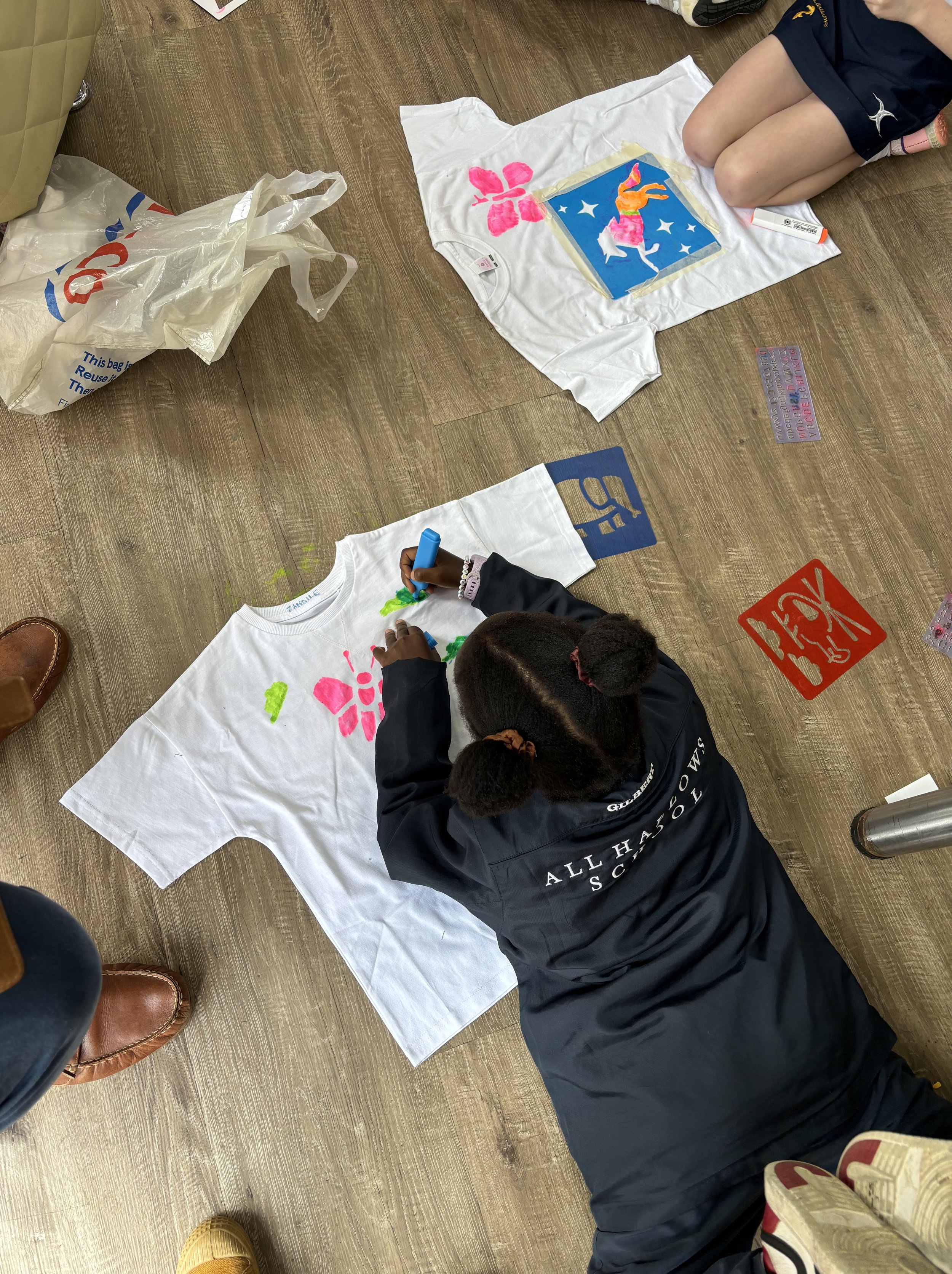 Child lying on the floor creating a colorful butterfly and flower design on a white T-shirt with fabric markers. Additional T-shirts with designs are on the floor nearby.