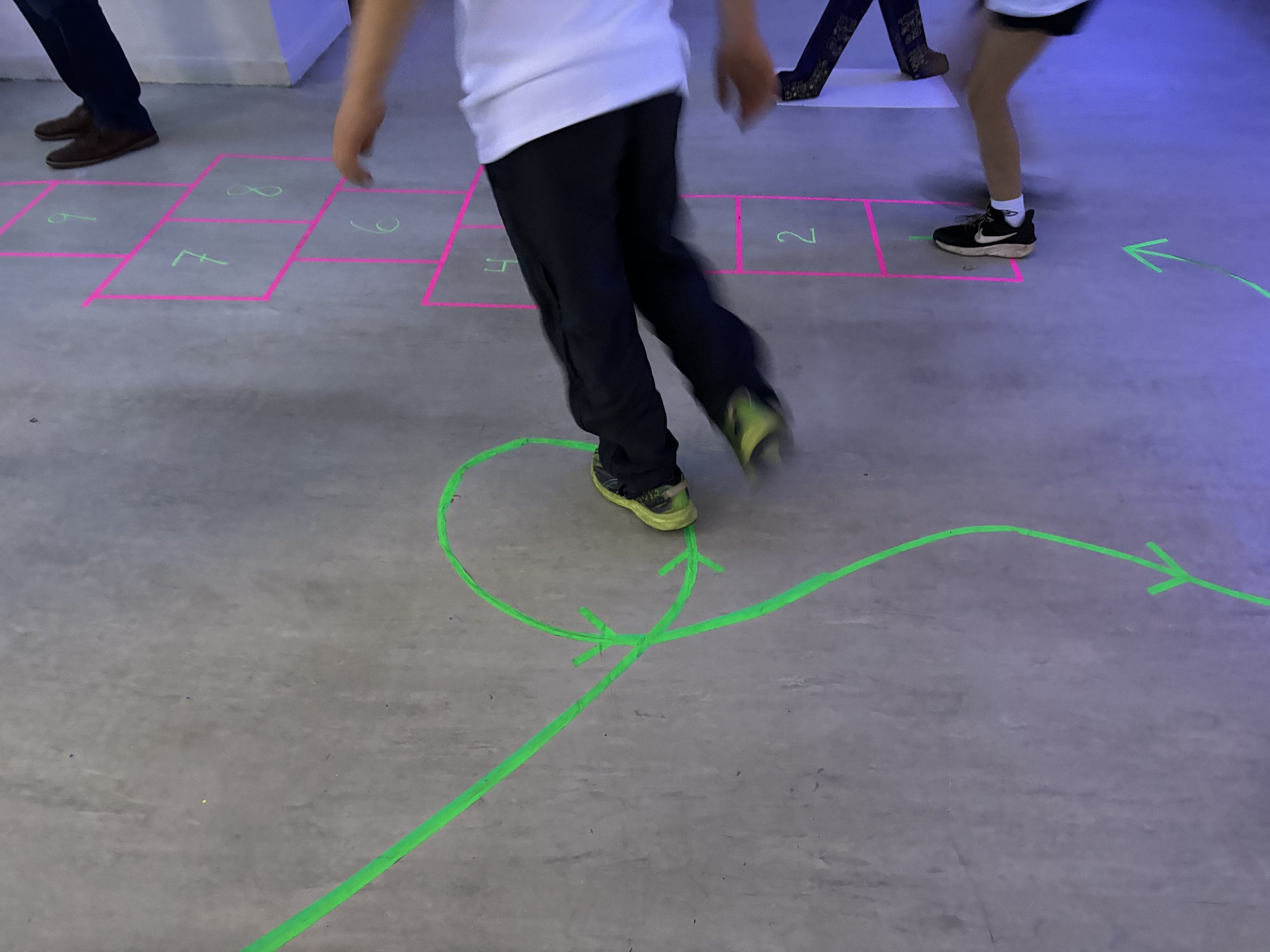 Children playing in a classroom with neon-colored hopscotch and hop directions drawn on the floor.