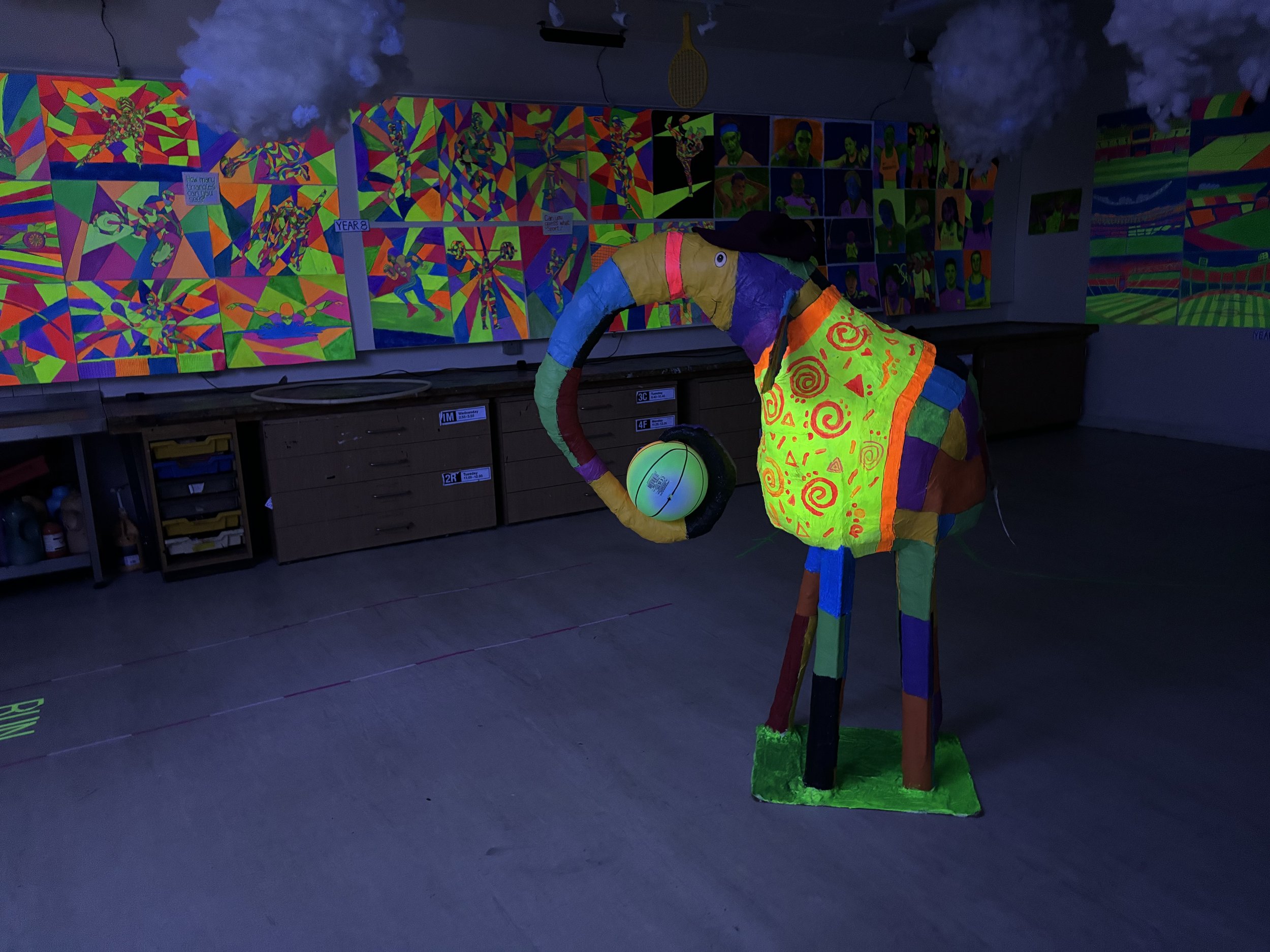 Colorful sculpture of a person holding a ball, decorated with bright patterns, in an art gallery with neon-colored artworks on the walls.