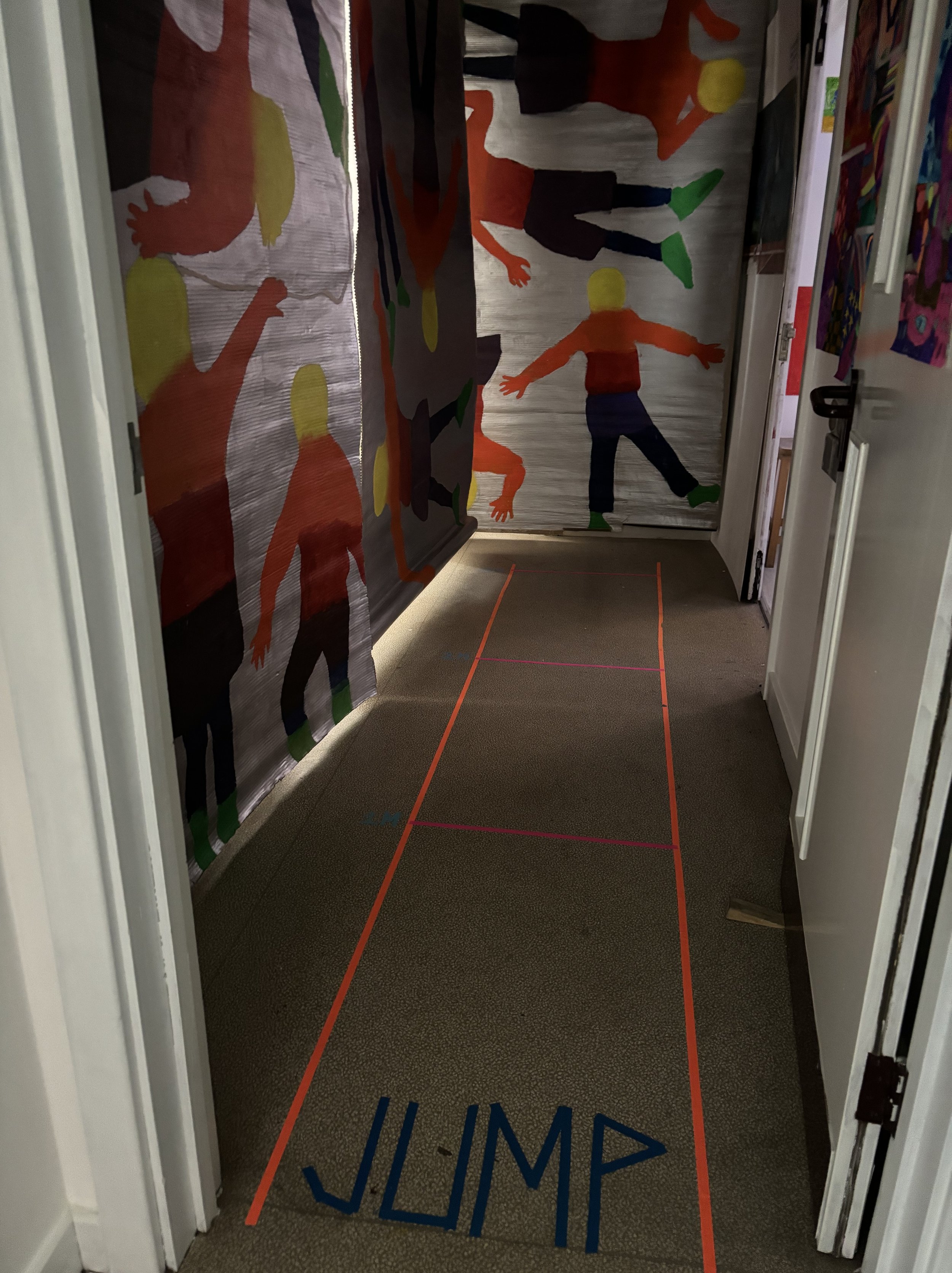 A hallway with colorful wall art featuring silhouettes of children playing, and orange tape on the floor outlining a hopscotch game.