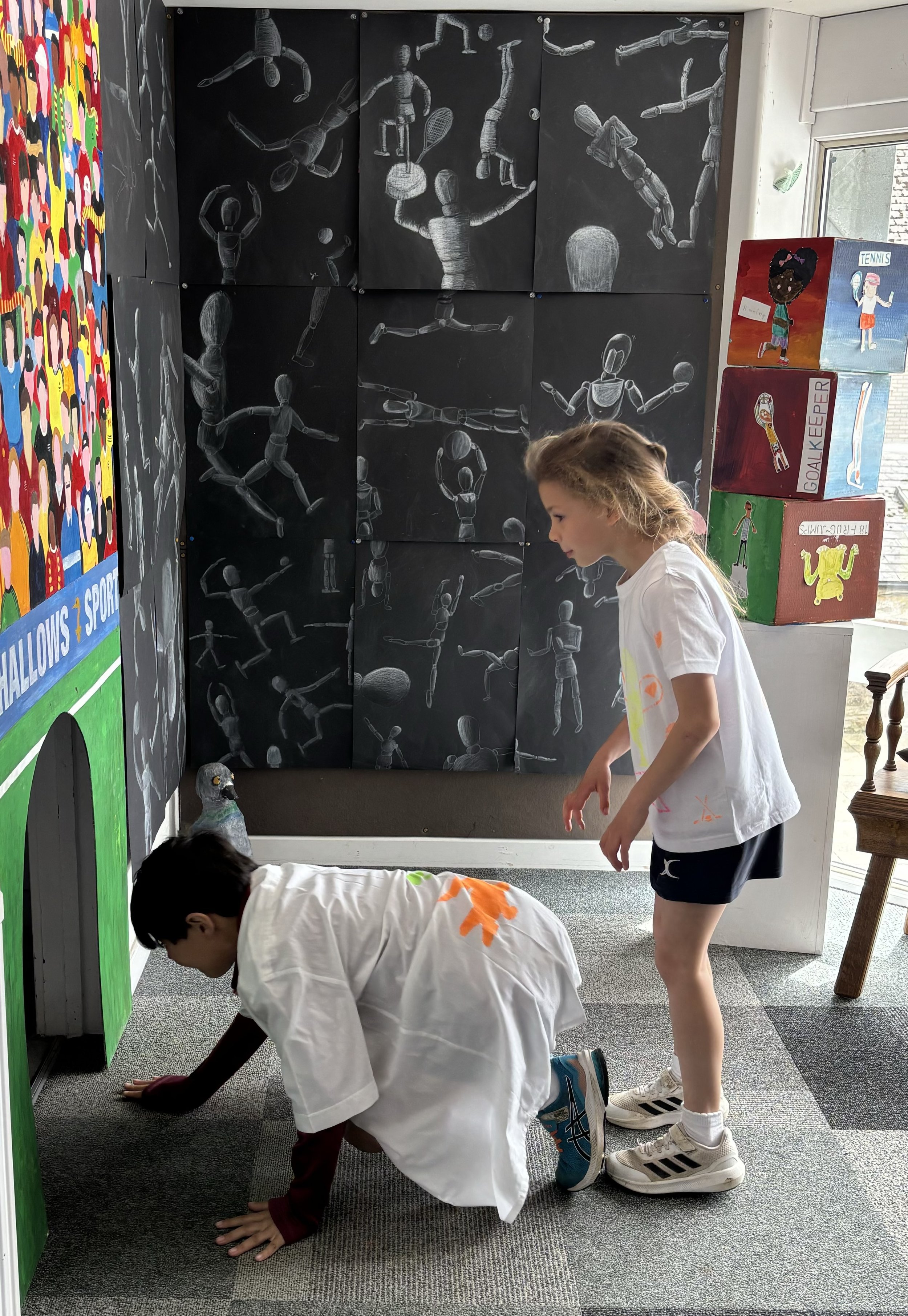 Two children, a girl and a boy, are playing indoors near a colorful wall with murals. The girl is standing, and the boy is on all fours. Behind them, there is a black chalkboard wall with drawings of human figures in various athletic poses, and a sta