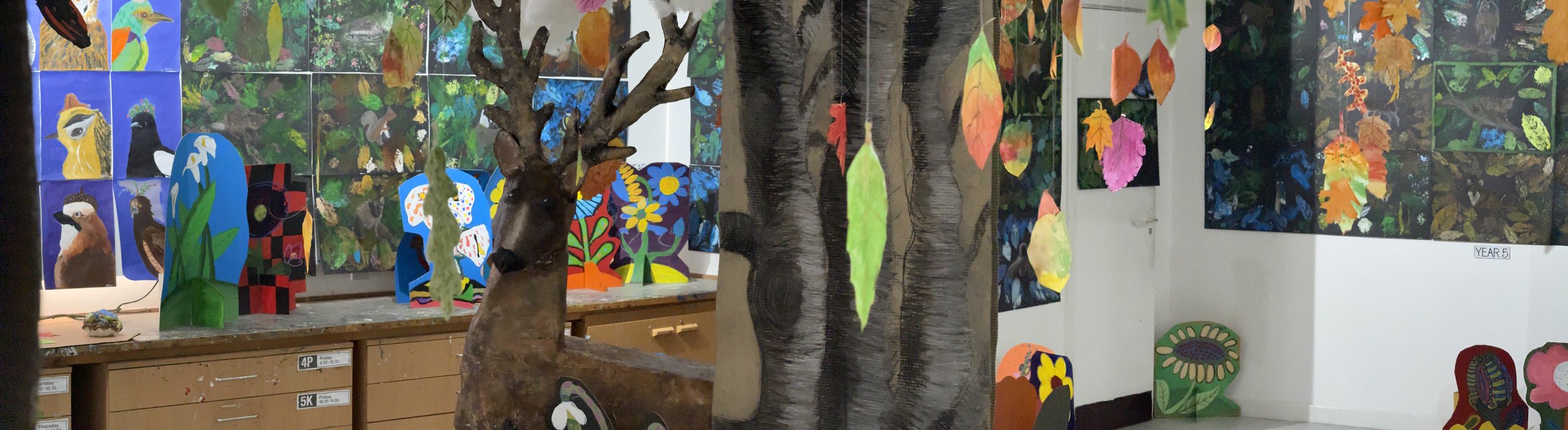Colorful art display in an art classroom, featuring paintings of animals and nature, cardboard trees, hanging leaf decorations, and painted cutouts.