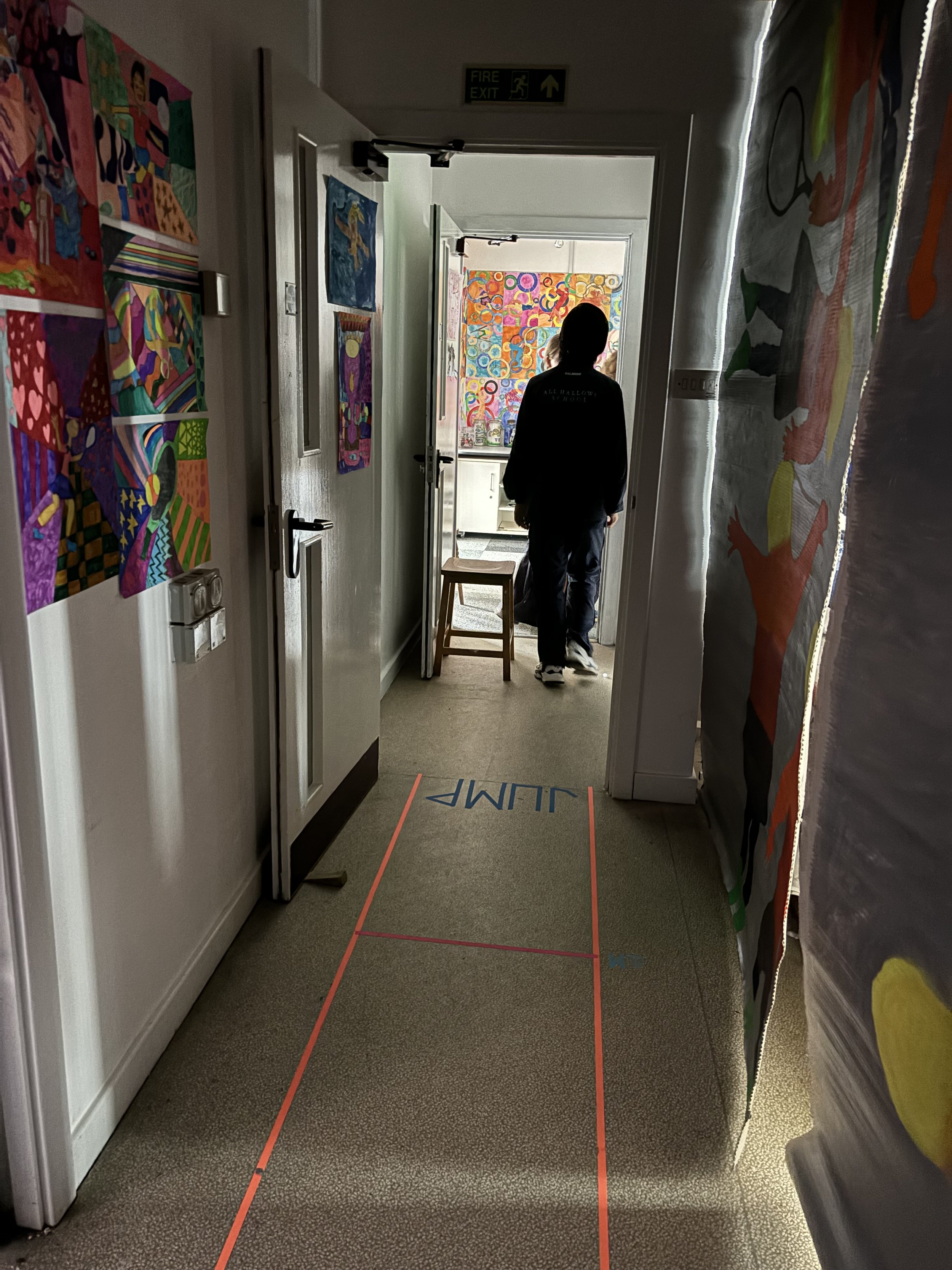 A hallway with colorful artwork on the walls, a person with dark hair walking towards a room with more vibrant art, and a floor marking labeled 'JUMP' with red tape lines.