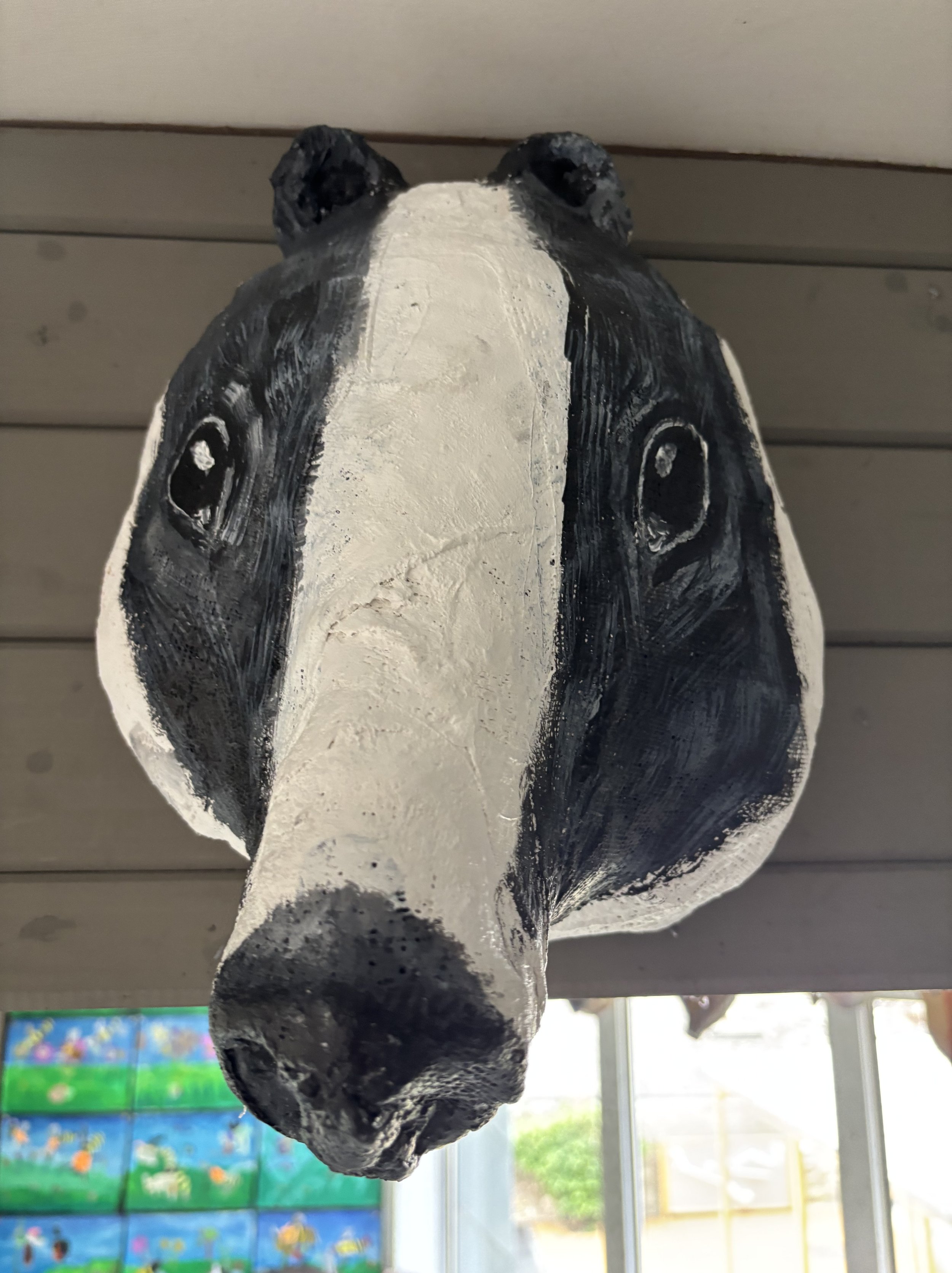 Handmade mask of a black and white badger’s face, viewed from below, mounted on a wall with horizontal wooden panels.