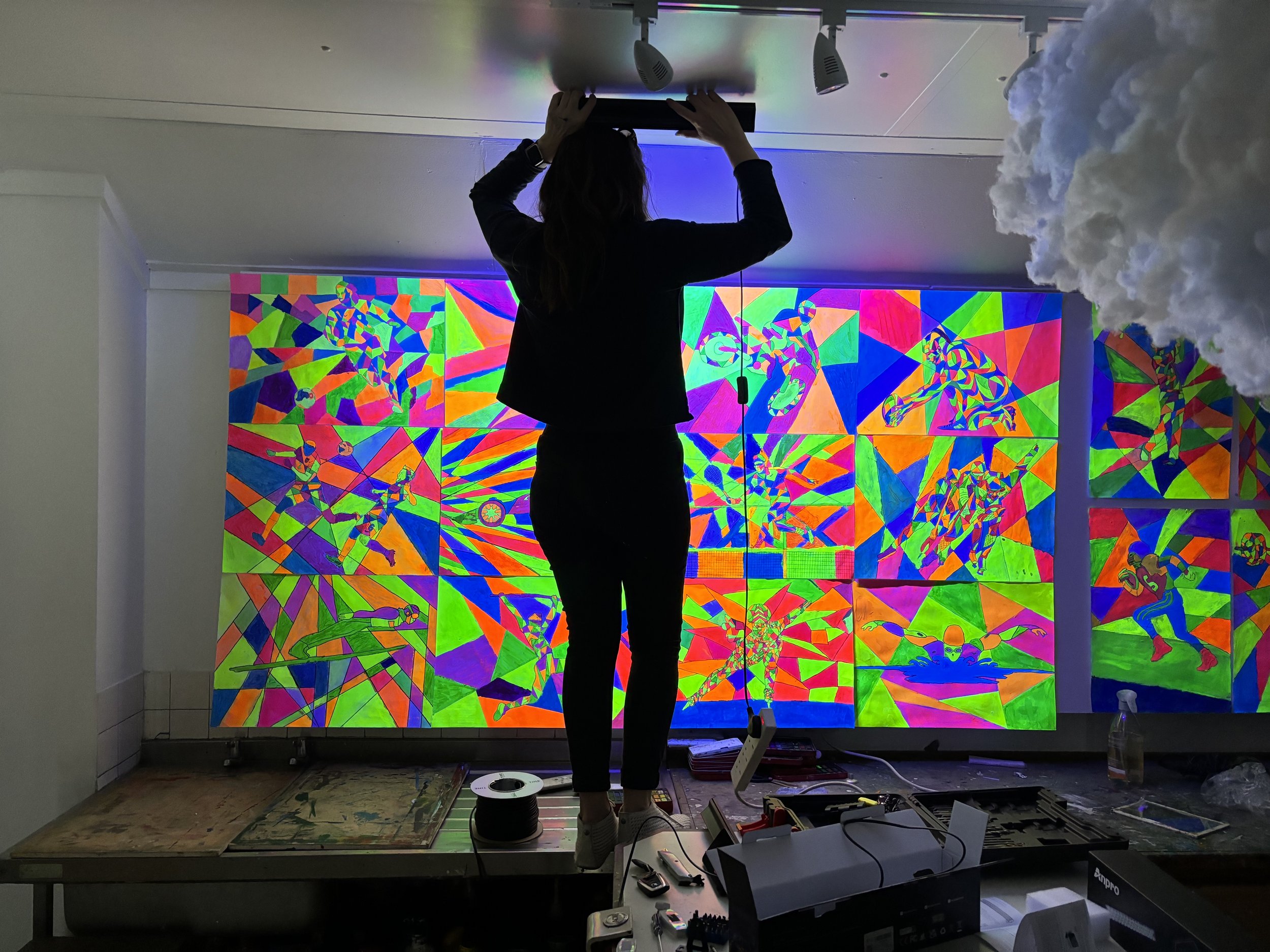 Person installing or adjusting a colorful, geometric stained glass artwork depicting athletes in motion, with various tools and equipment on a table in front.
