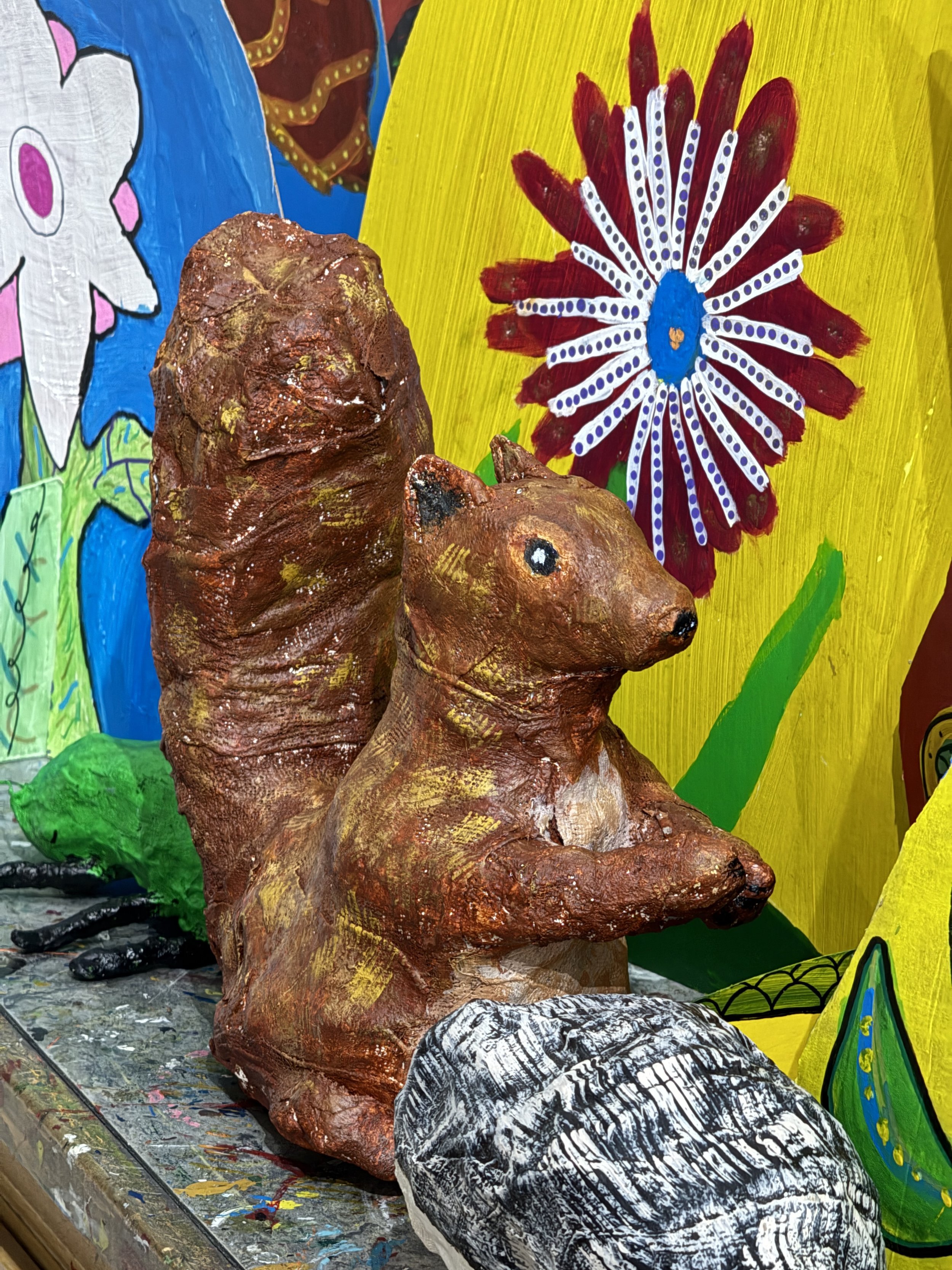 Painted art sculpture of a squirrel with a bushy tail, surrounded by colorful, abstract painted backgrounds and objects.