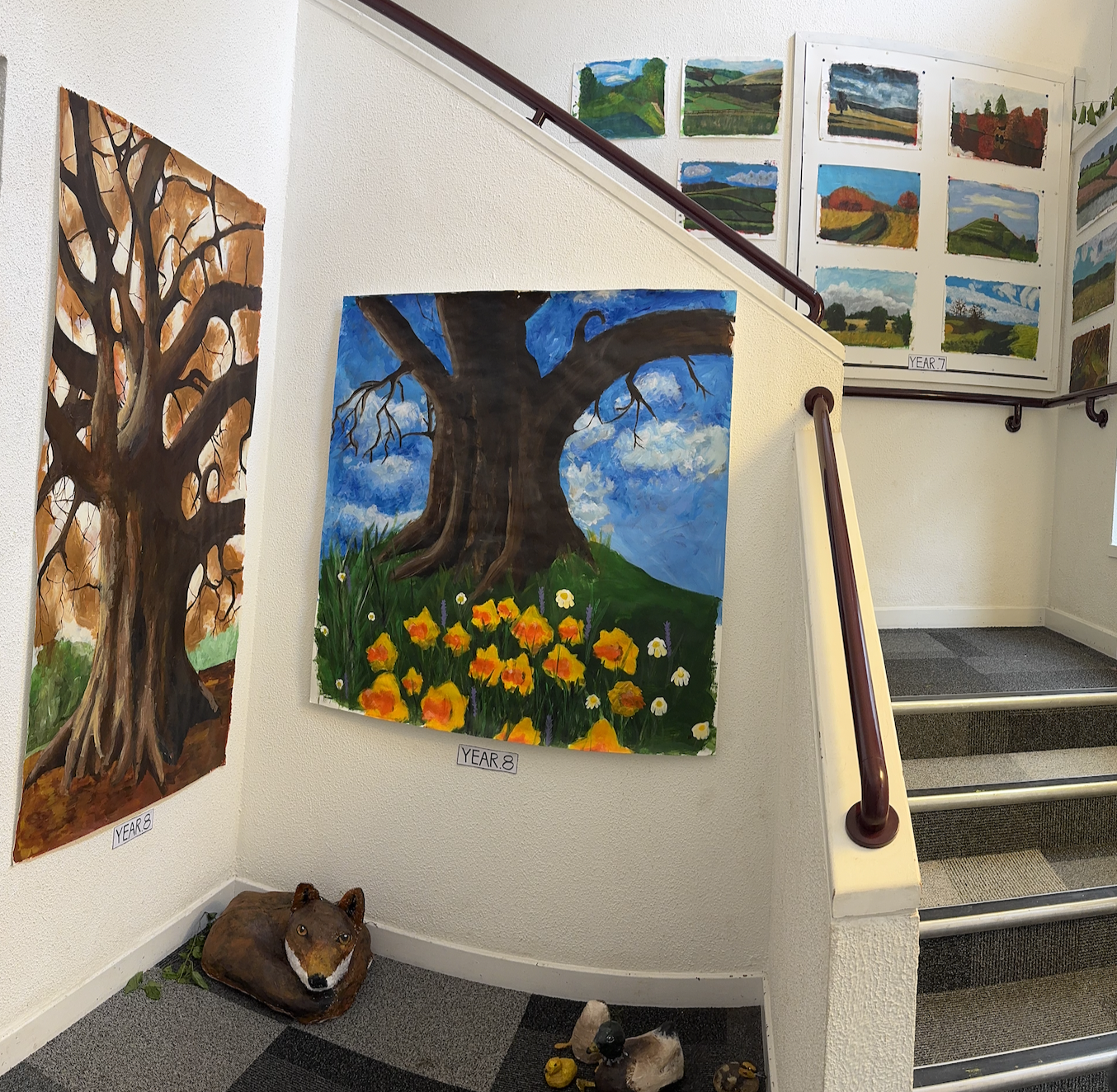 Art display on a staircase featuring painted landscape and nature scenes, including a large tree with flowers at its base, and animal sculptures on the floor.