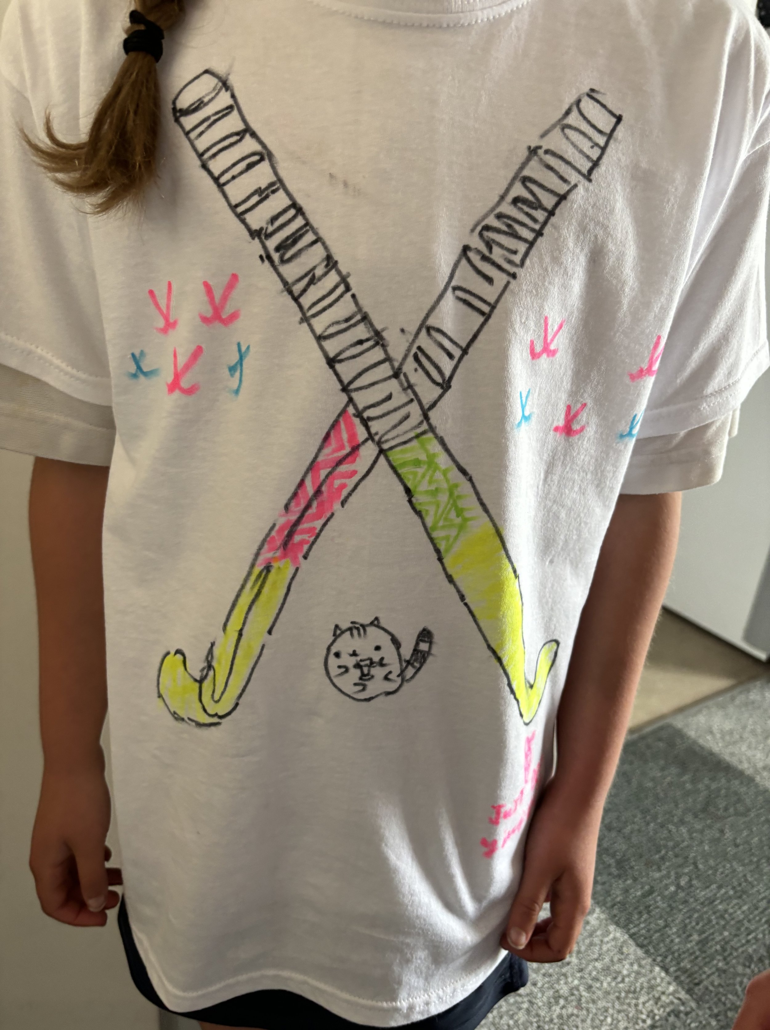 A white T-shirt with a colorful hand-drawn design of two crossed hockey sticks, a small cat, and pink and blue flying birds.