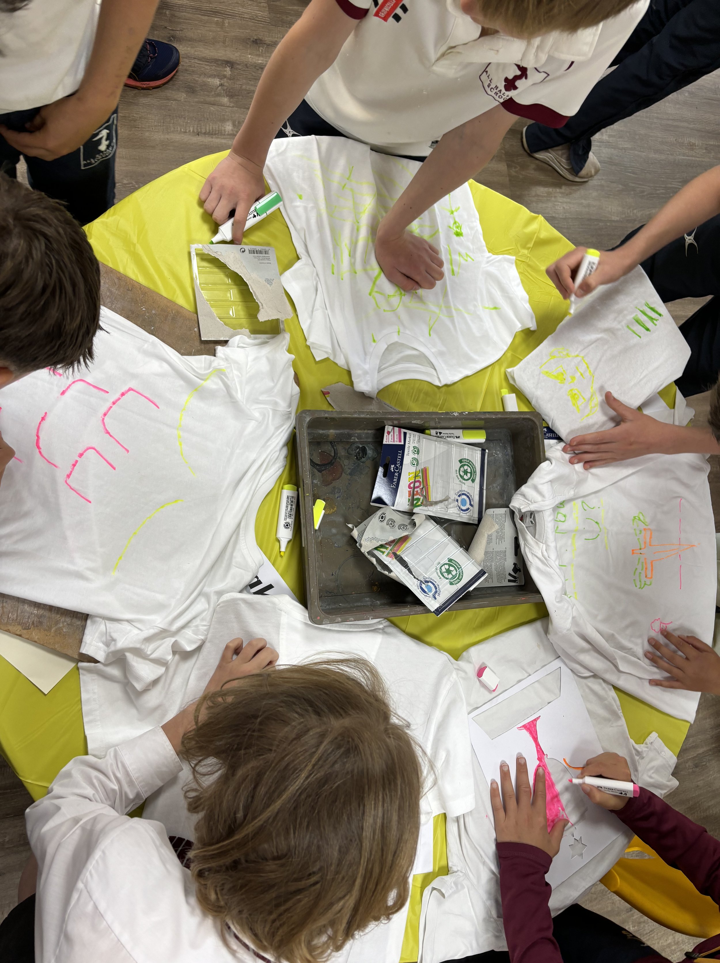 Children gathered around a table, decorating white T-shirts with colorful markers for a craft activity.