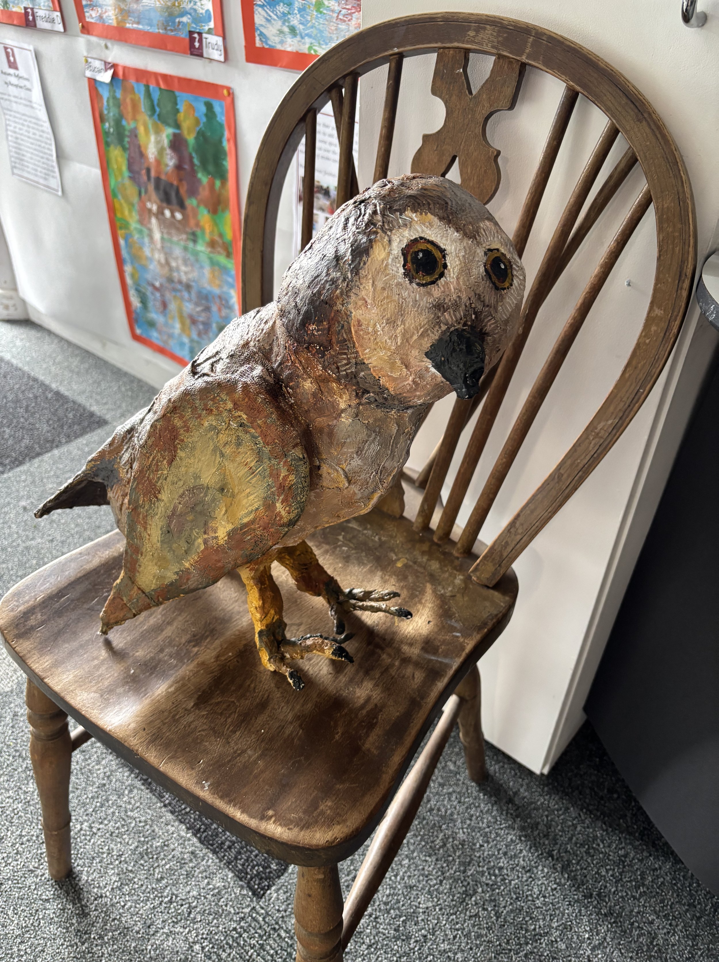 A handmade paper mache sculpture of a bird sitting on a wooden chair with a slatted backrest. The bird has painted features, large eyes, and a beak, with textured, multi-colored feathers.