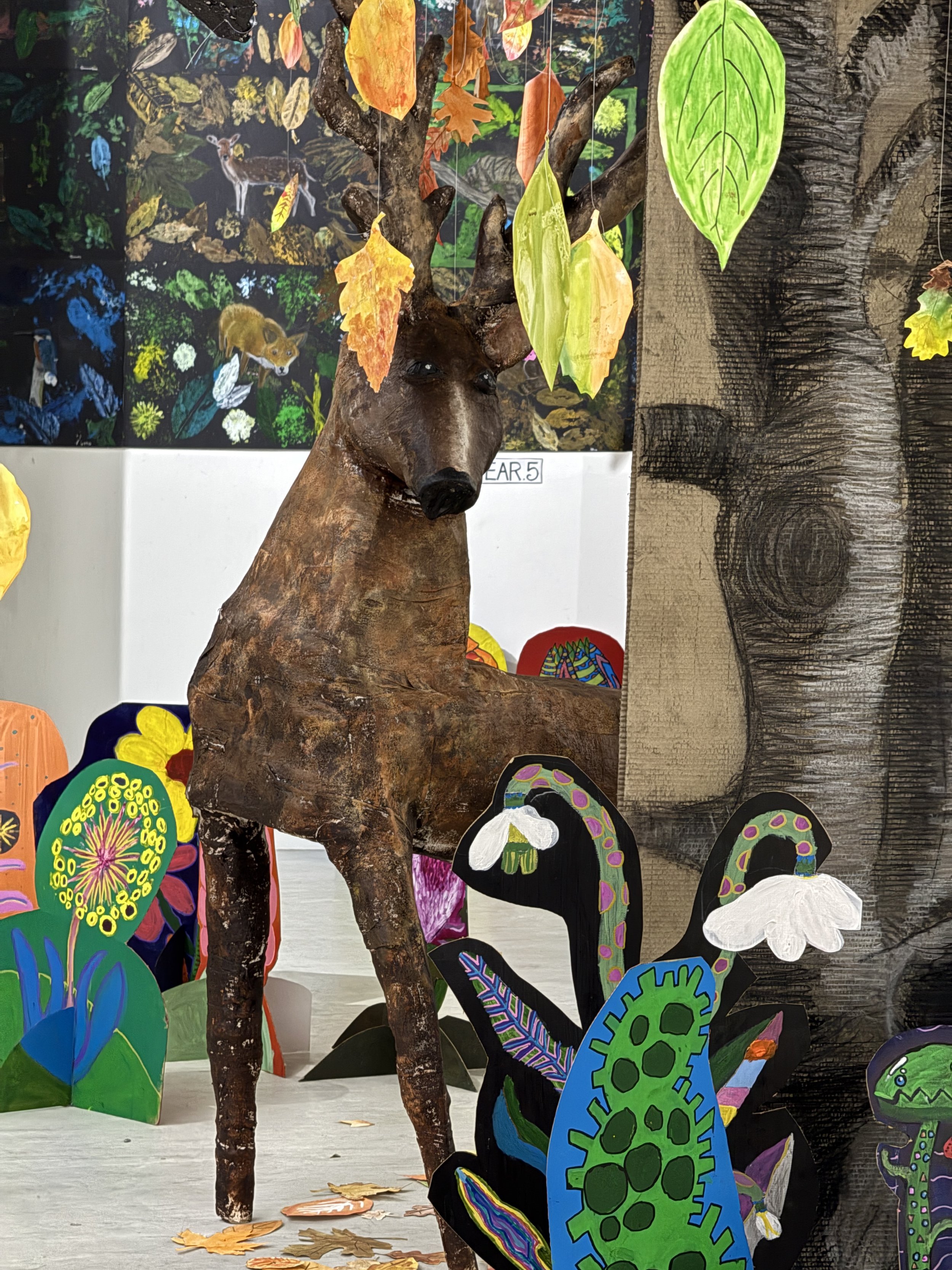 A craft project featuring a sculpture of a deer with antlers, surrounded by a tree trunk and colourful leaves attached, set against a background of painted paper plants and forest-themed artwork.