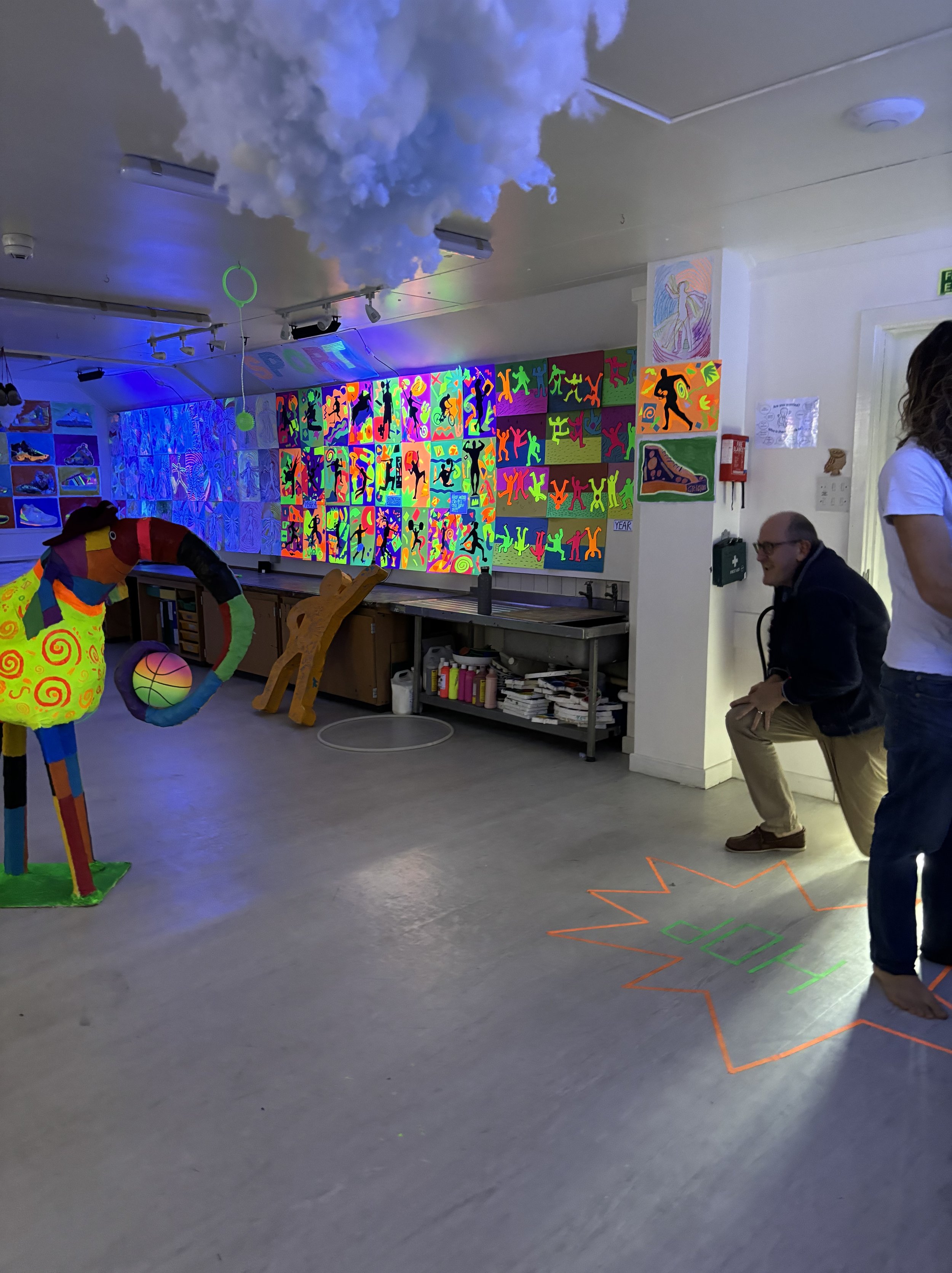 Indoor art space with colorful paintings and sculptures, neon lighting, and people engaging in a game of hopscotch on the floor.