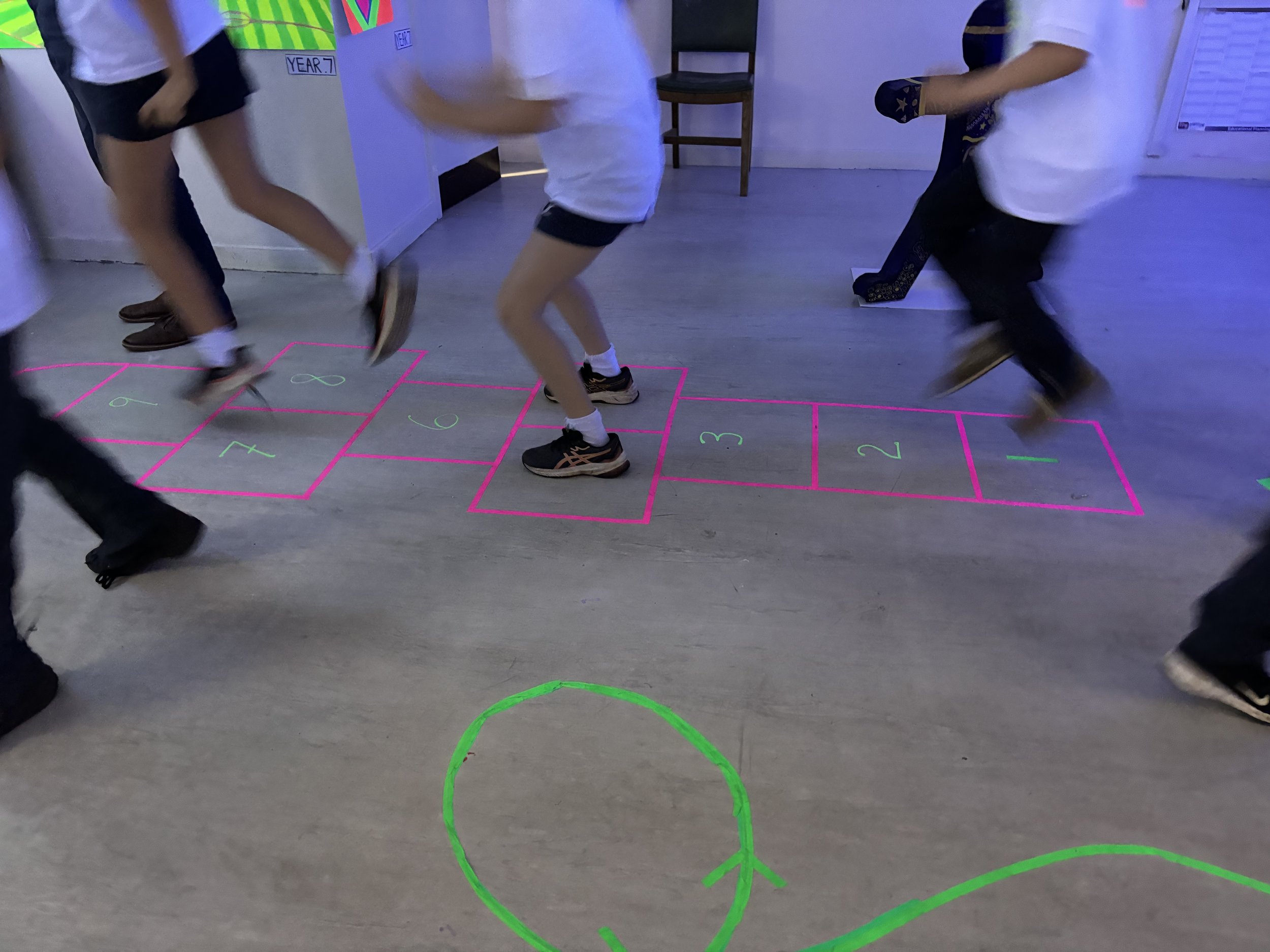 Children playing hopscotch on a floor with neon-colored chalk outline. Some children are mid-jump, and others are waiting their turn.