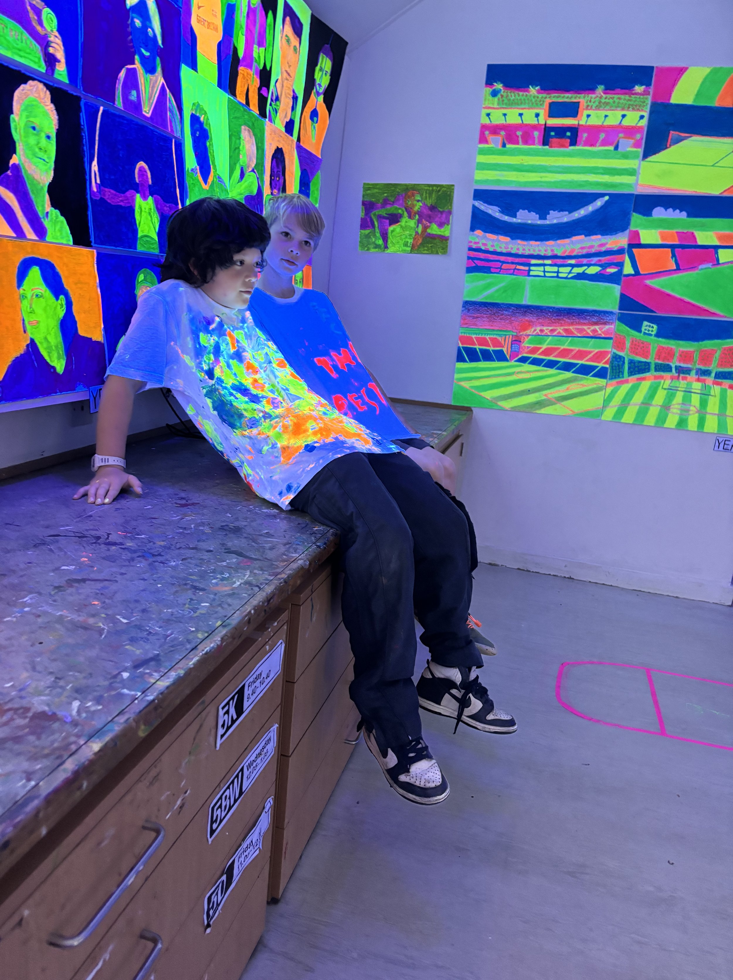 Two boys sitting on a wooden table in an art gallery with glow-in-the-dark paintings on the walls, including portraits and sports stadium scenes.