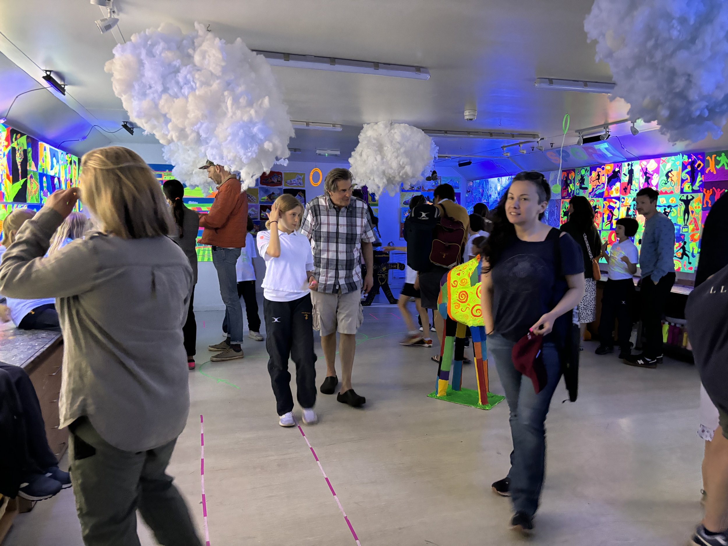 People in an indoor art exhibit with colorful, illuminated artwork and cloud-themed decorations hanging from the ceiling.