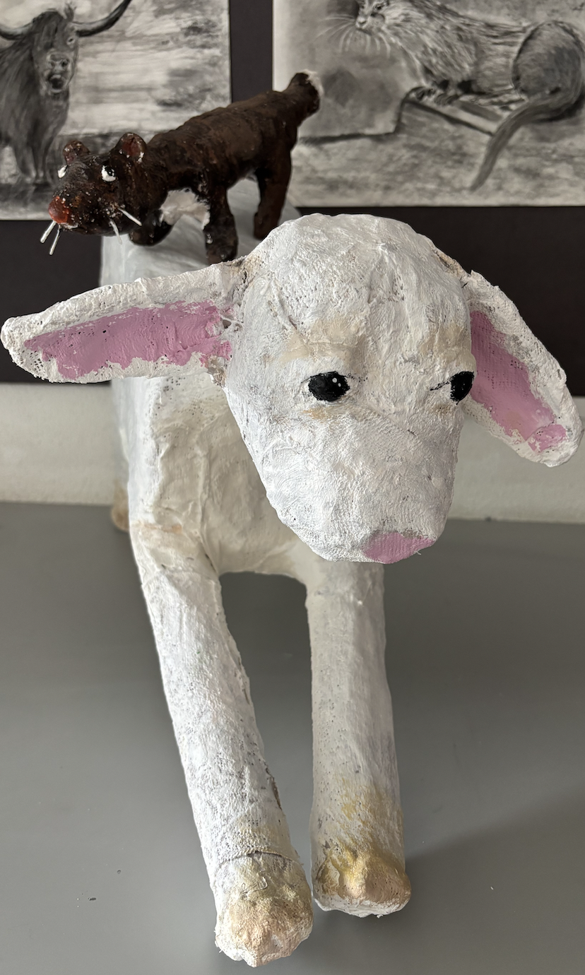 A white, handmade sculpture of a dog with pink on the inside of its ears and a pink nose, with a brown, homemade animal figurine resembling a rat or squirrel placed on its back.