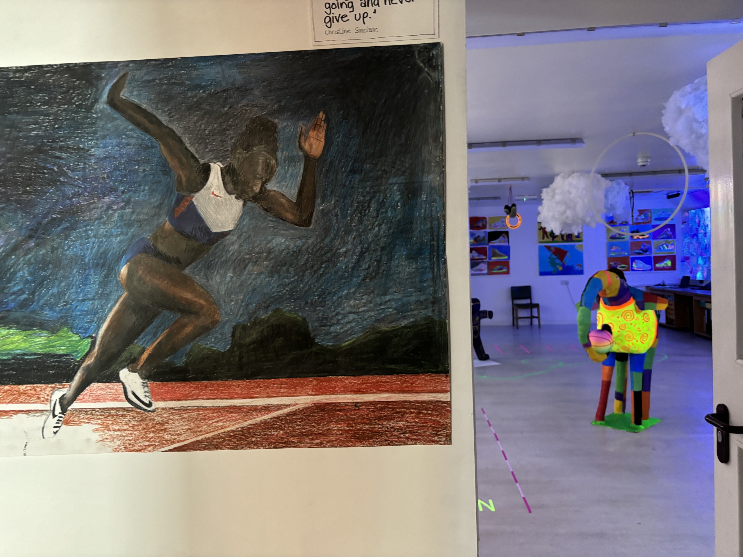 A painting of a female sprinter starting a race on a track with a dark sky background. To the right, through an open door, a colorful indoor space with artwork on the walls and cloud-shaped decorations hanging from the ceiling can be seen.