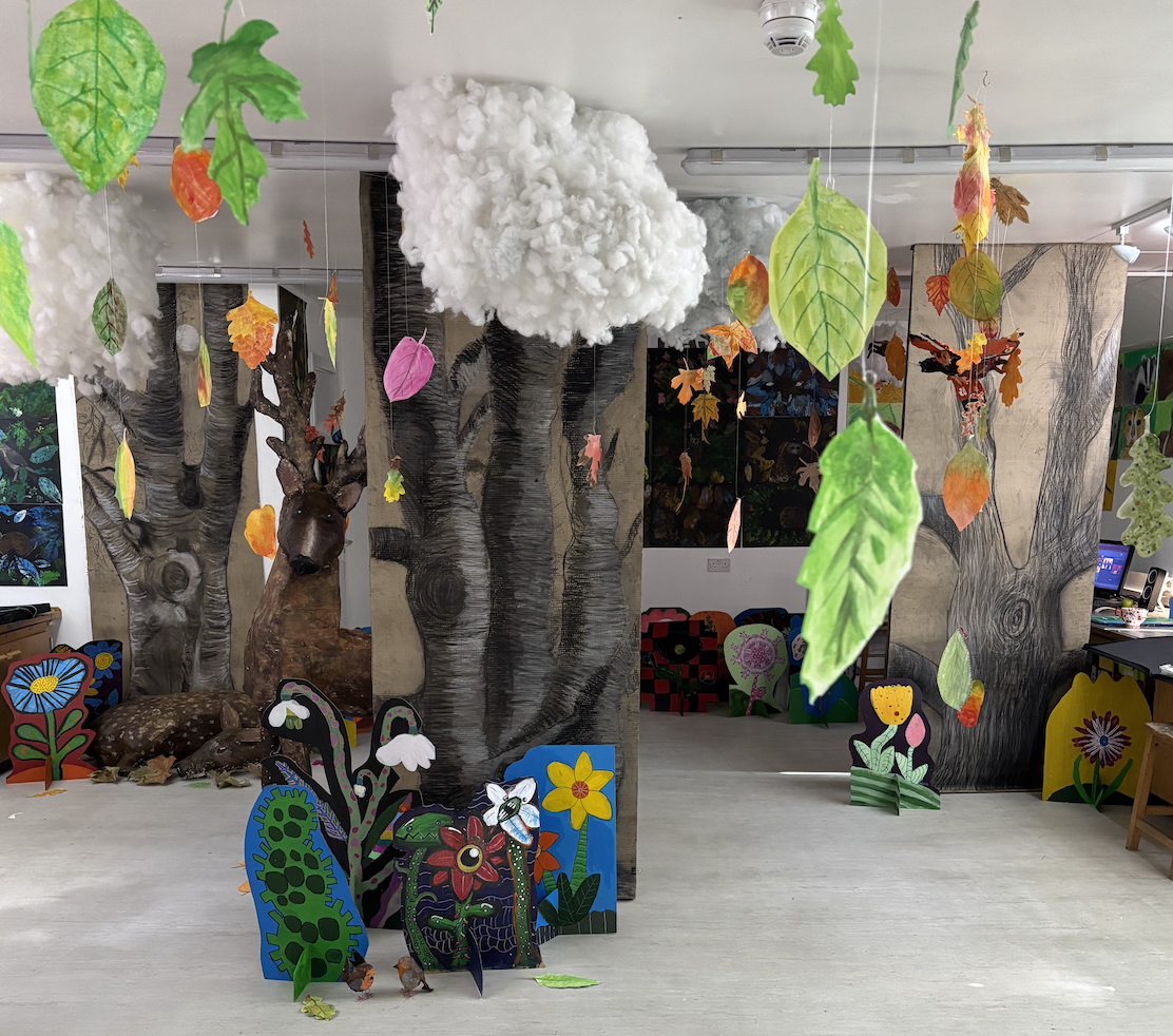 Interior of a colorful children's play or art area with paper lanterns resembling clouds and hanging leaves, painted trees, animal sculptures, and decorative cutouts of flowers and insects.